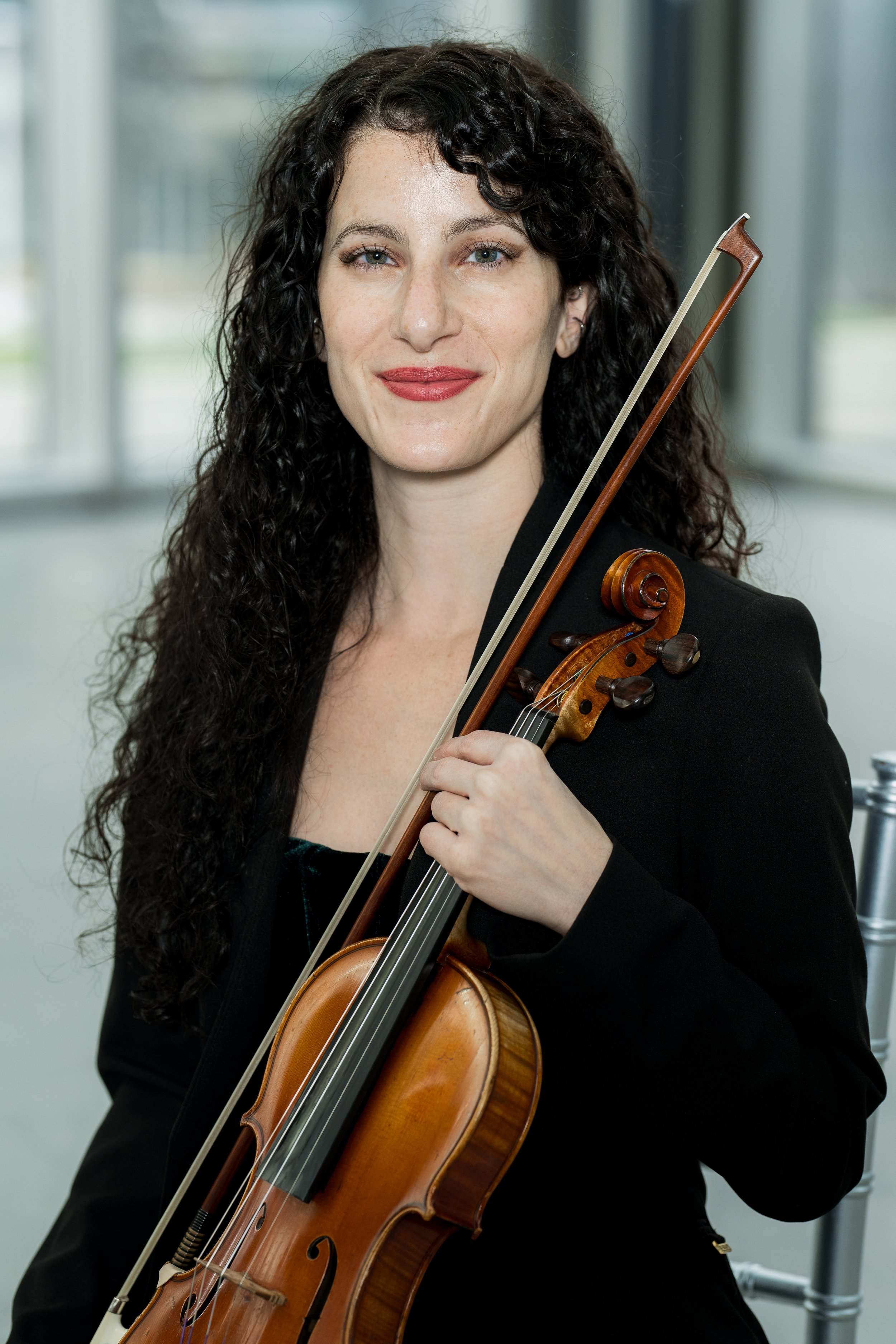 Cara Garofalo holding a violin and a bow, wearing a black blazer, in a bright indoor setting.