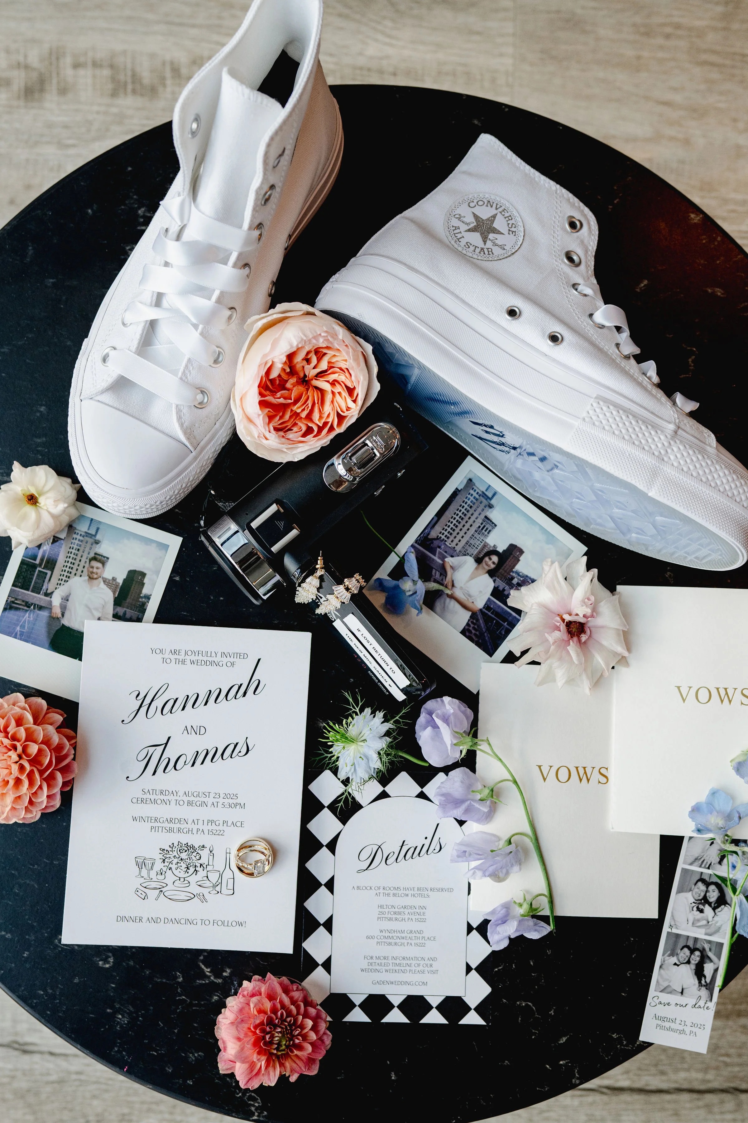 White Converse sneakers, pink and white flowers, wedding invitation for Hannah and Thomas, wedding details card, wedding vows booklet, wedding photos, wedding rings, lipstick, nail polish, and a video camera on a black round table.
