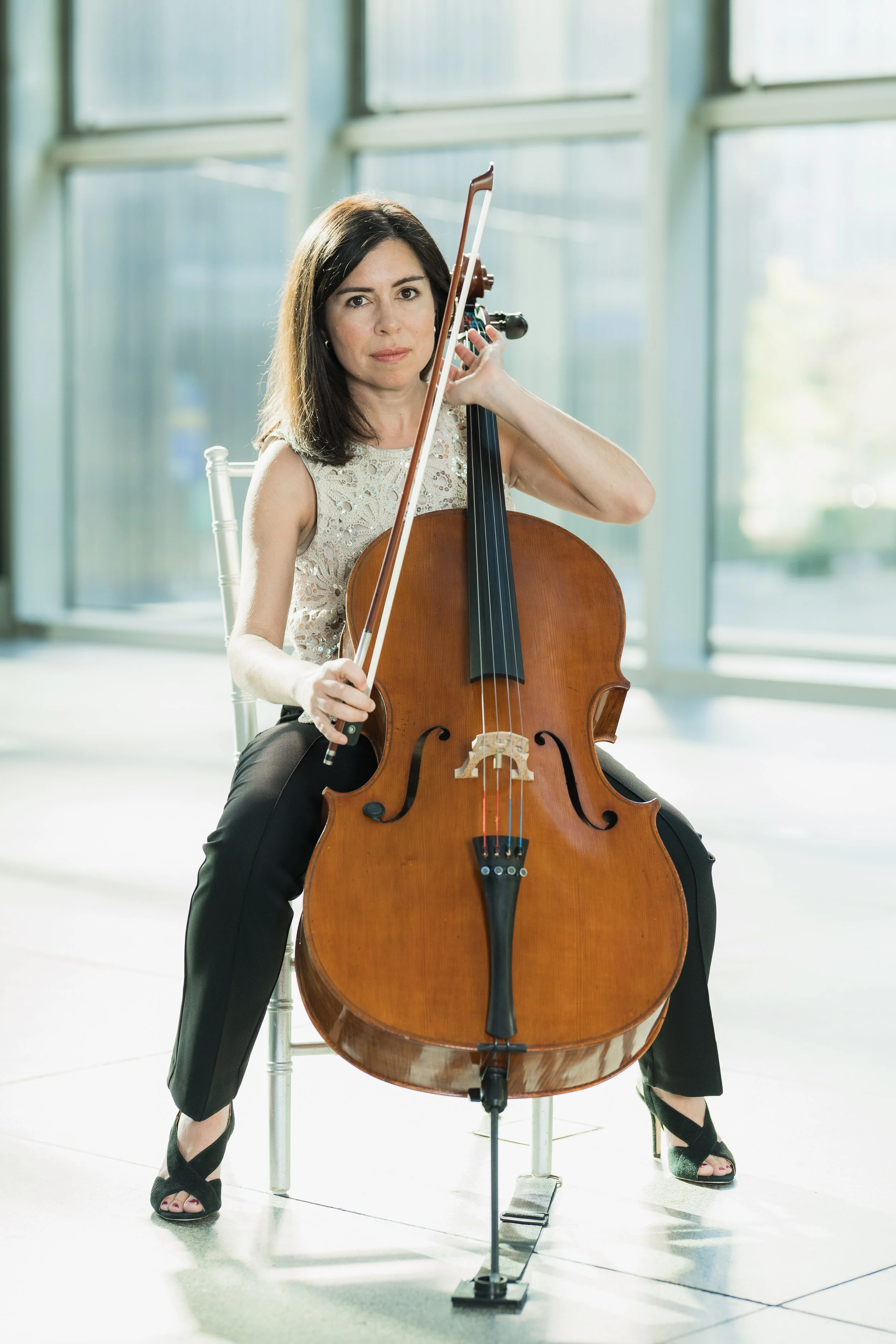 Becky Keeley playing a cello indoors, sitting on a chair, with large windows in the background.