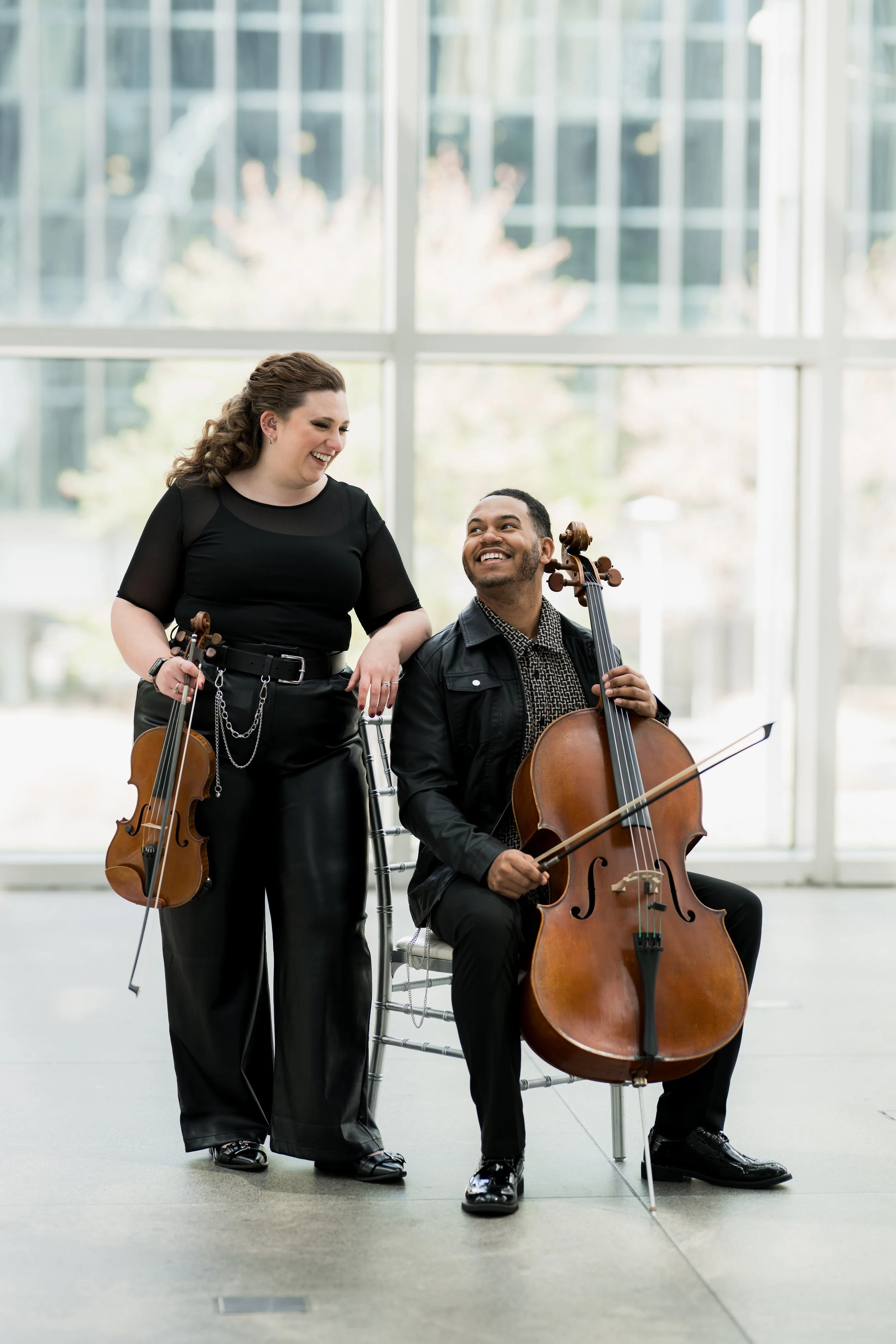 Two musicians, Nicole Lennartz holding a violin and Ahmer'e Blackman sitting with a cello, smiling at each other inside a space with large windows.