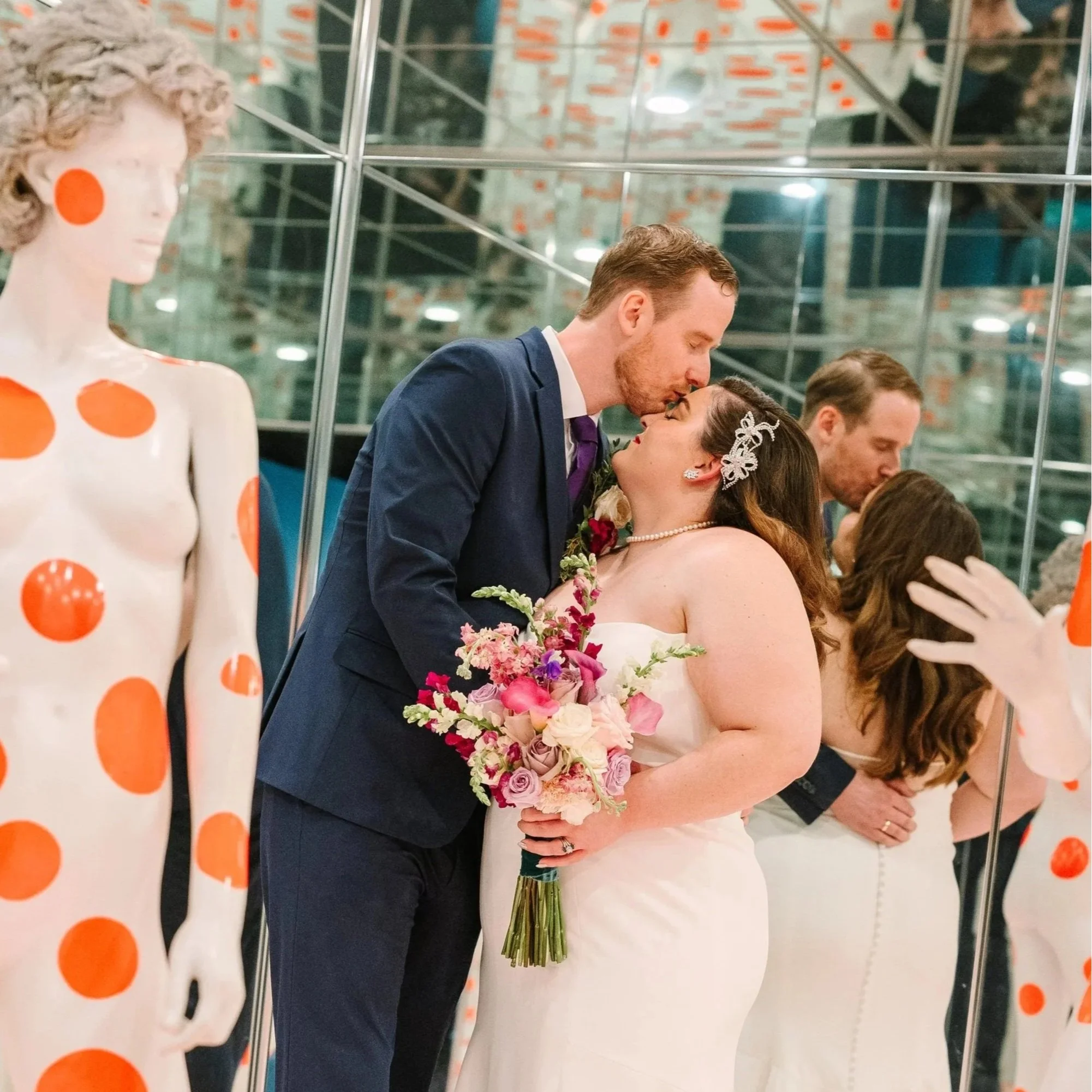 Devan and Jamie shares a kiss on their wedding day surrounded by mirrors and mannequins with orange polka dots at the Mattress Factory.