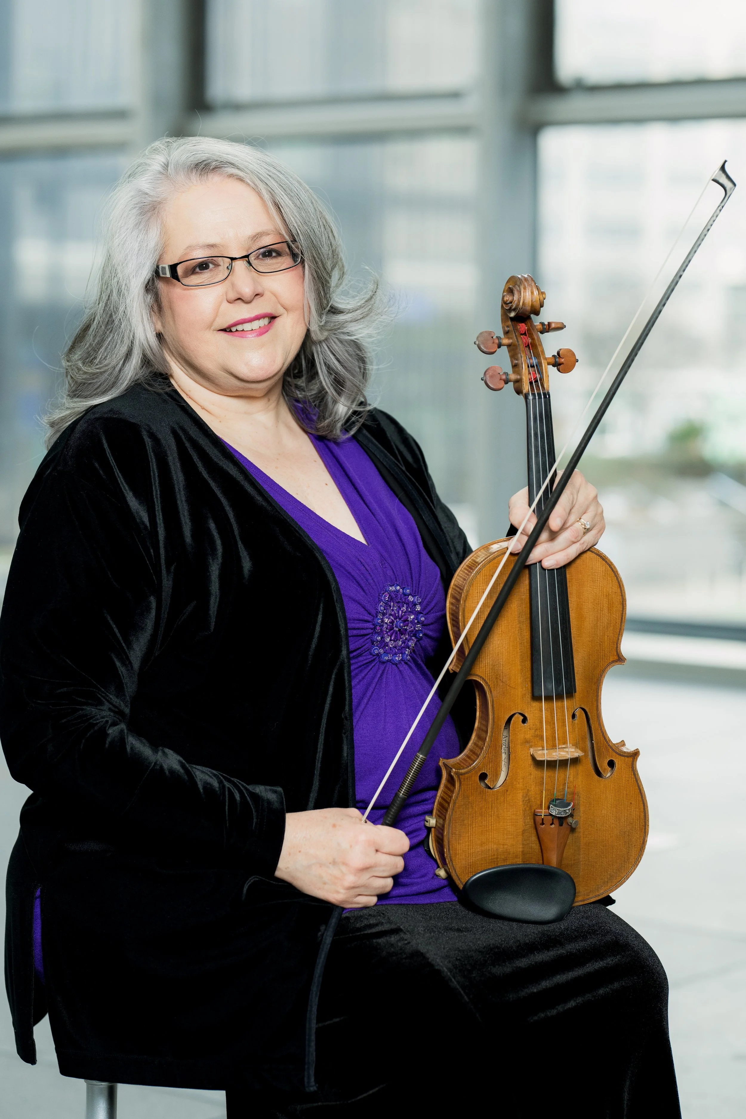 Jennifer Madge smiling while holding a violin and bow. She is seated indoors with large windows in the background.