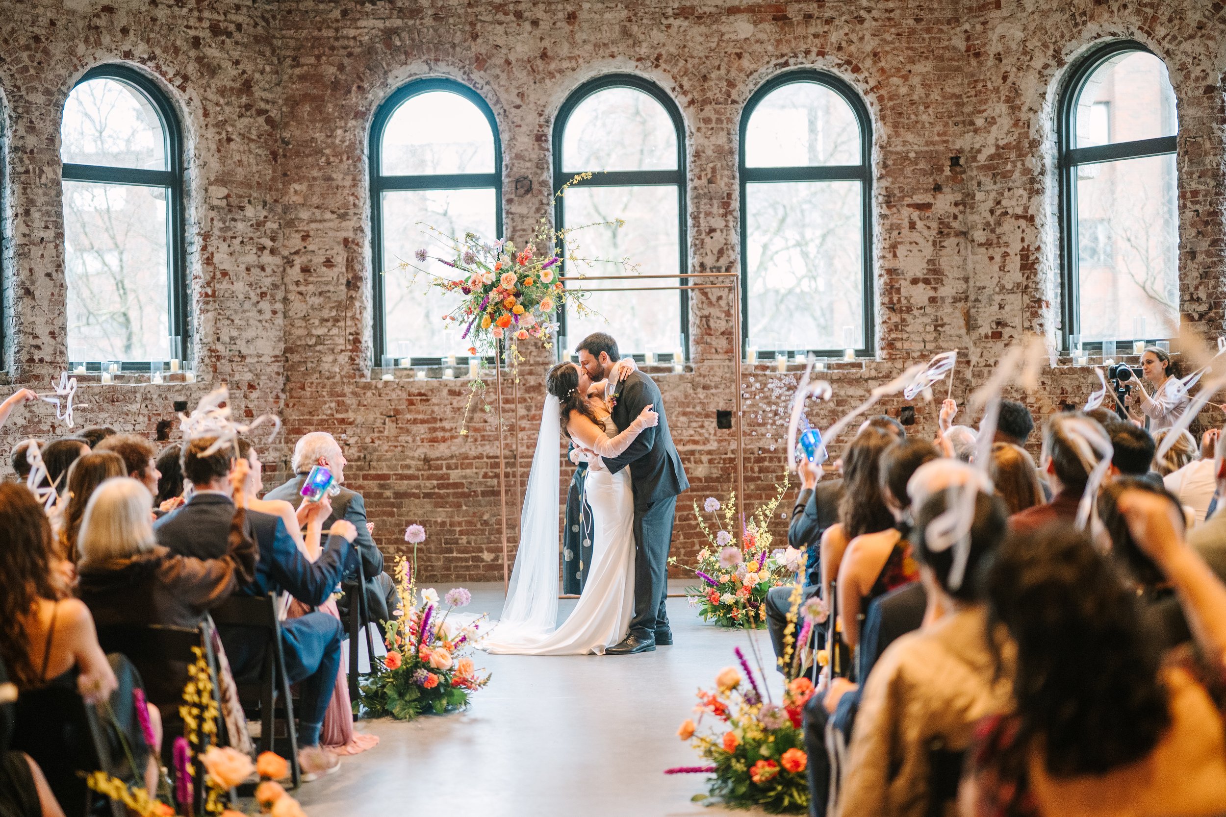 A wedding ceremony inside a rustic brick building with large arched windows. The bride and groom are sharing a kiss beneath a floral arch, surrounded by seated guests celebrating with confetti and flowers.