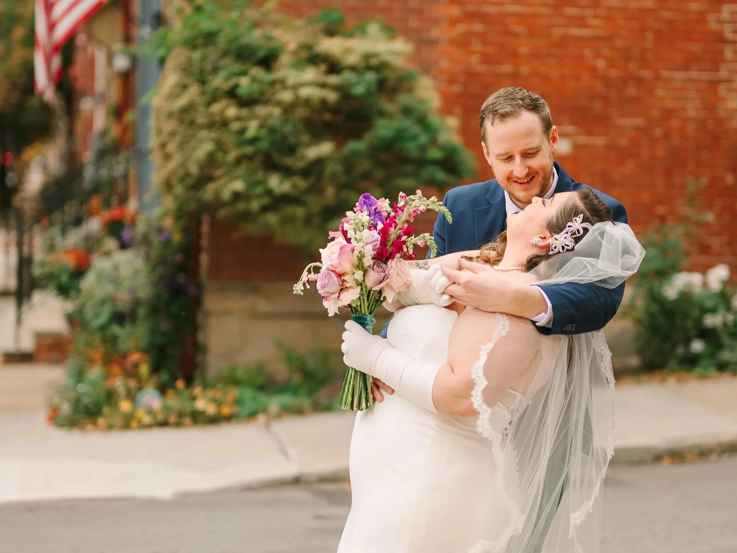 Devan and Jamie in their wedding attire outside the Mattress Factory. Devan is smiling and holding a bouquet of flowers as Devan holds onto her.