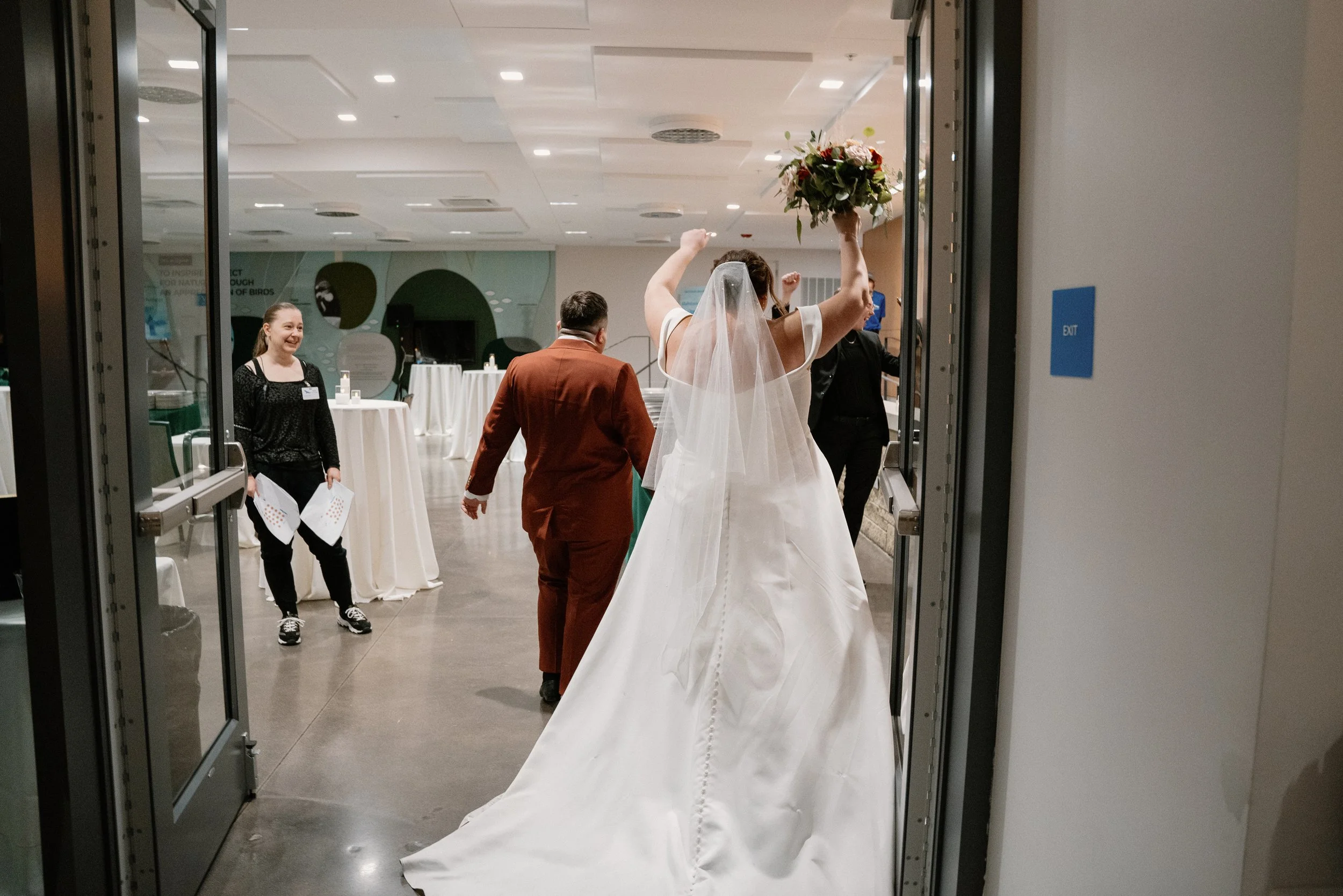 Bride with a bouquet celebrating as she enters a reception hall, entering through glass doors, wearing a white wedding gown and veil.