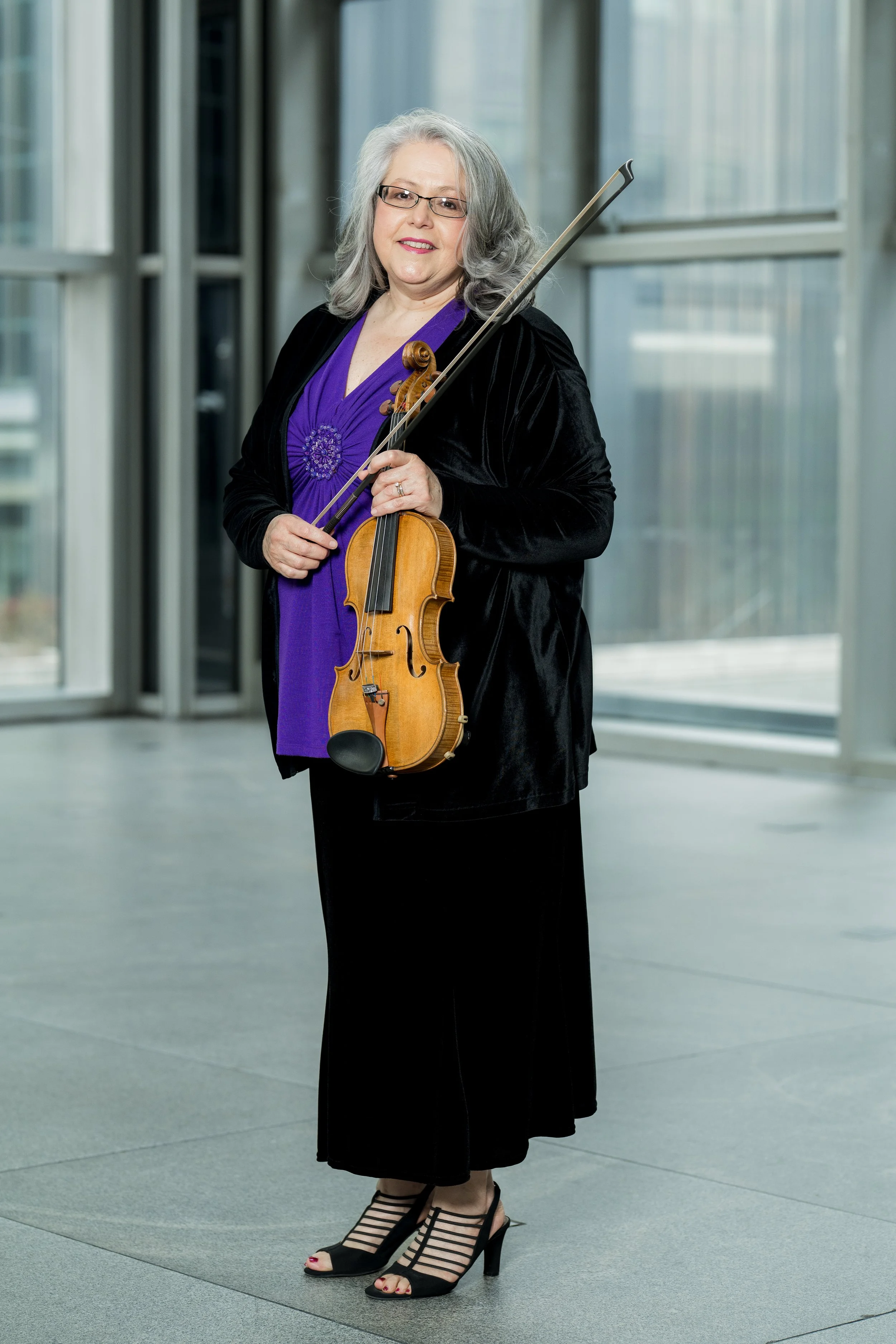 Jennifer Madge holding a violin and bow inside a modern building with large glass windows.