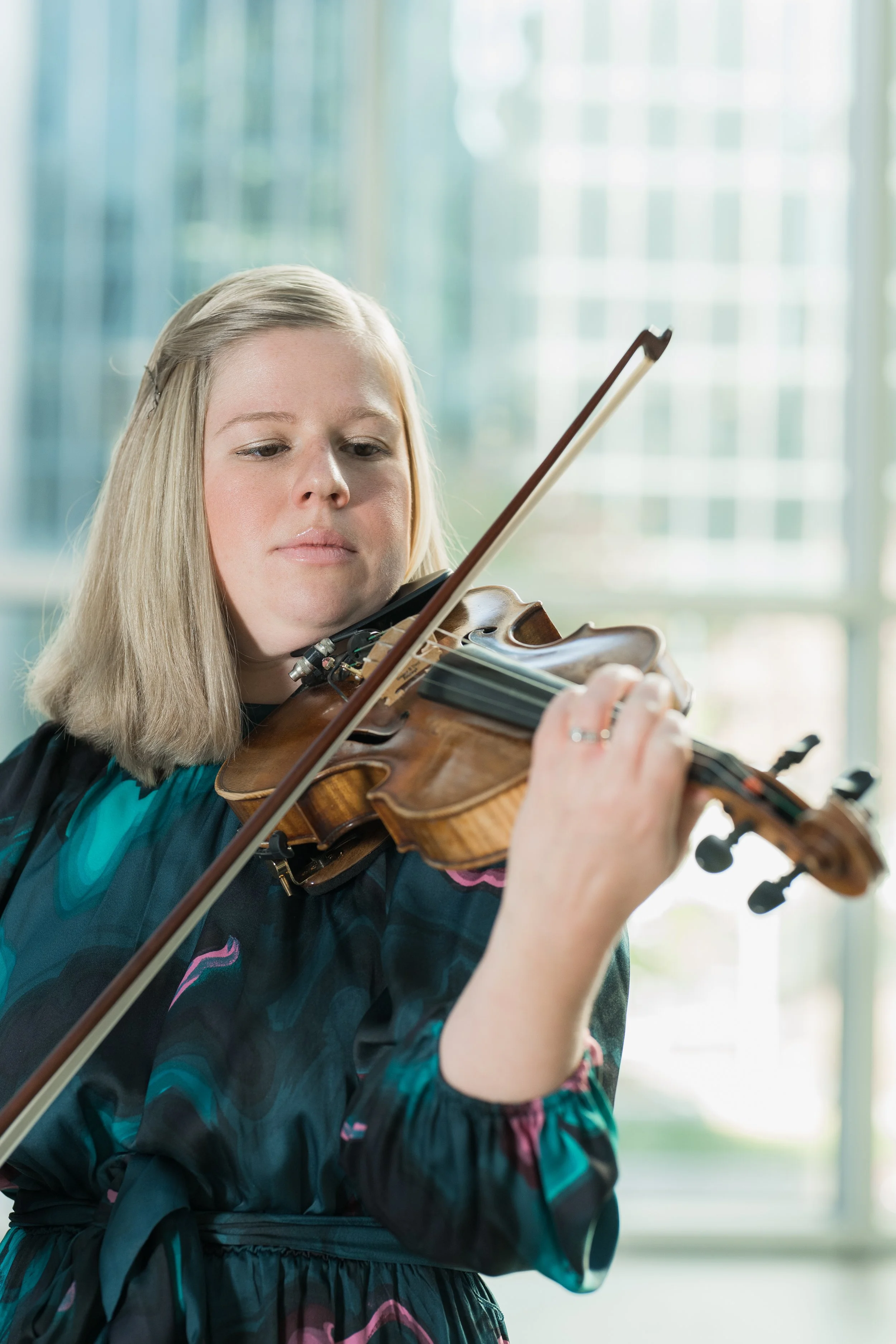 Amber Hooke playing her violin indoors with natural light coming through large windows.