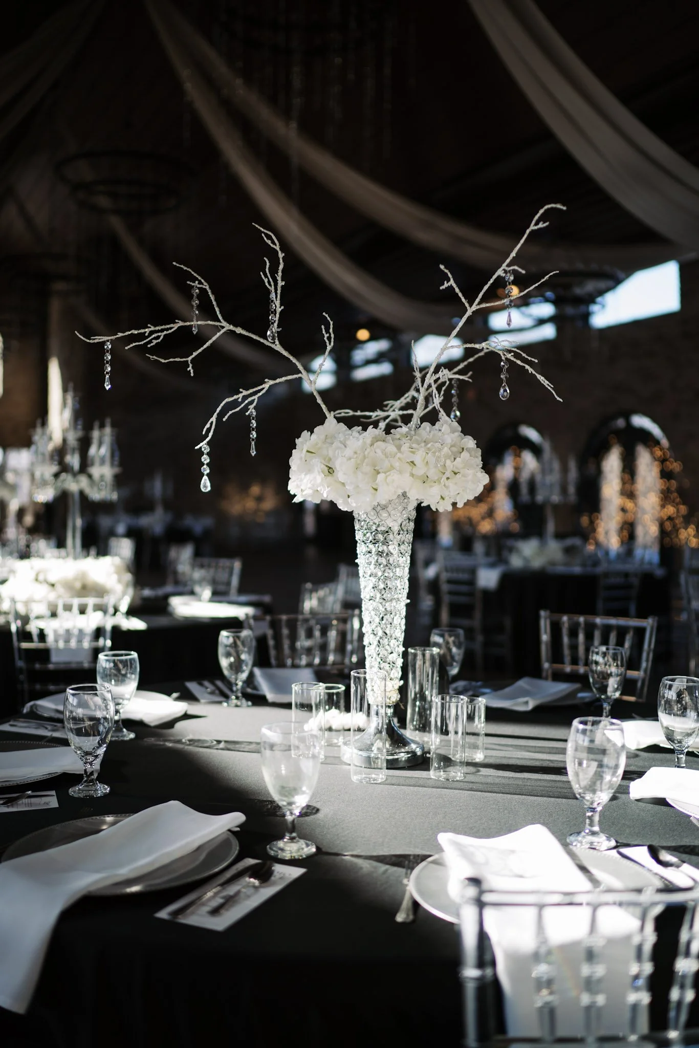 Elegant event table with black tablecloth, white napkins, multiple glasses, and a tall crystal vase with white flowers and hanging crystal decorations at Bella Amore Enchanted Acres.