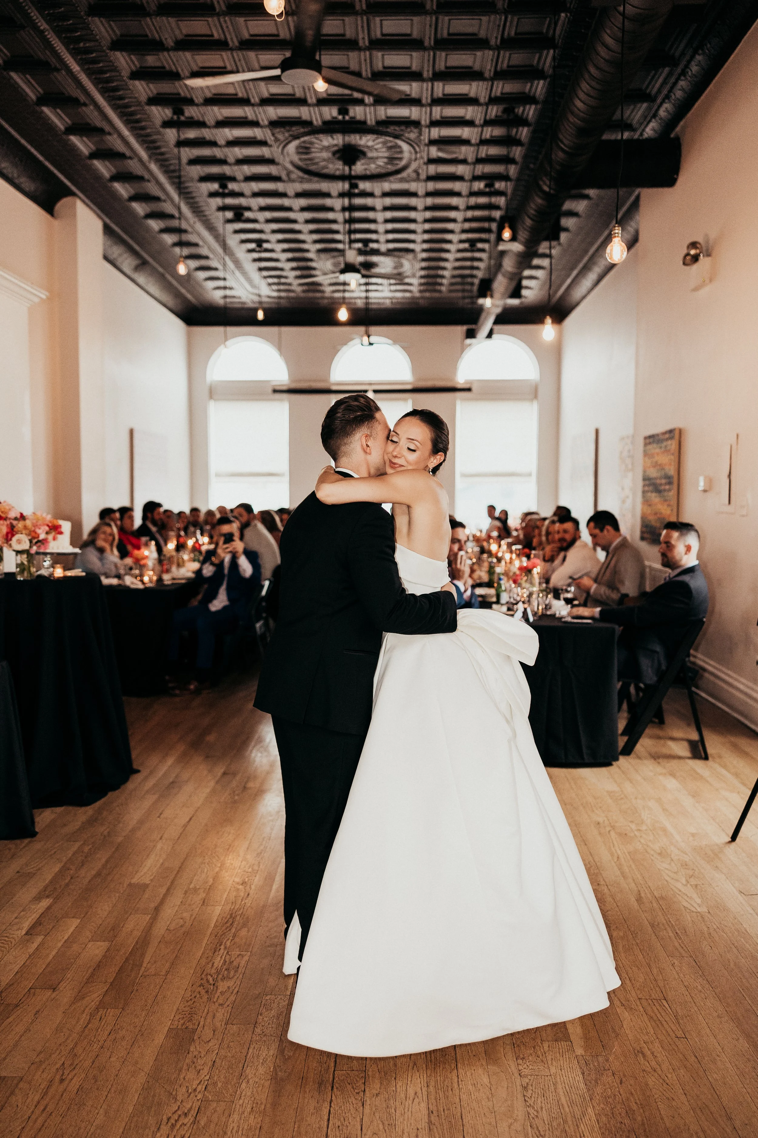 Bride and groom sharing their first dance at a wedding reception in a decorated venue with seated guests watching.