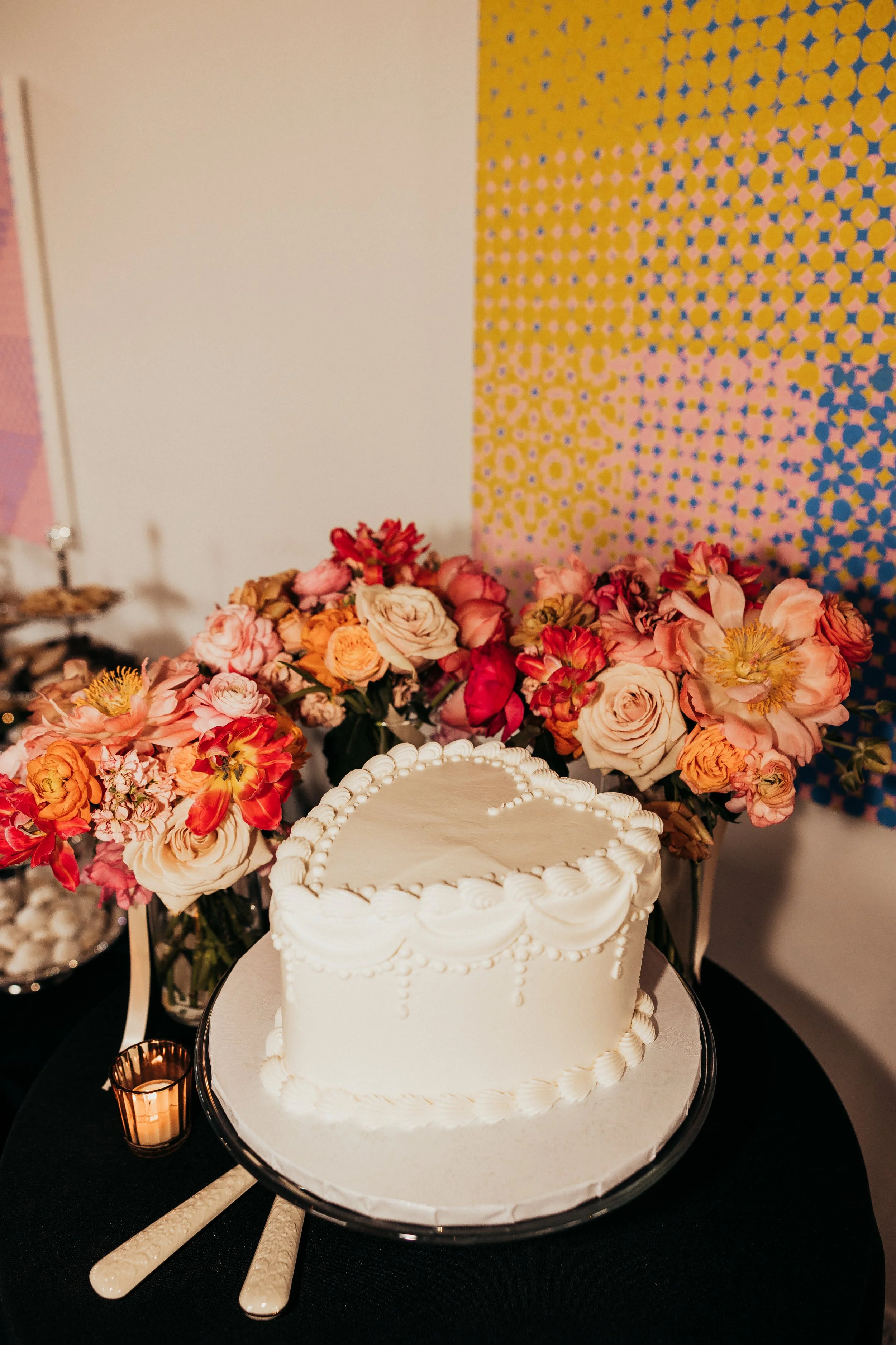 A white heart-shaped cake with piped frosting on a round silver tray, surrounded by pink and orange flowers, candles, and dessert utensils on a black table with colorful geometric art in the background.
