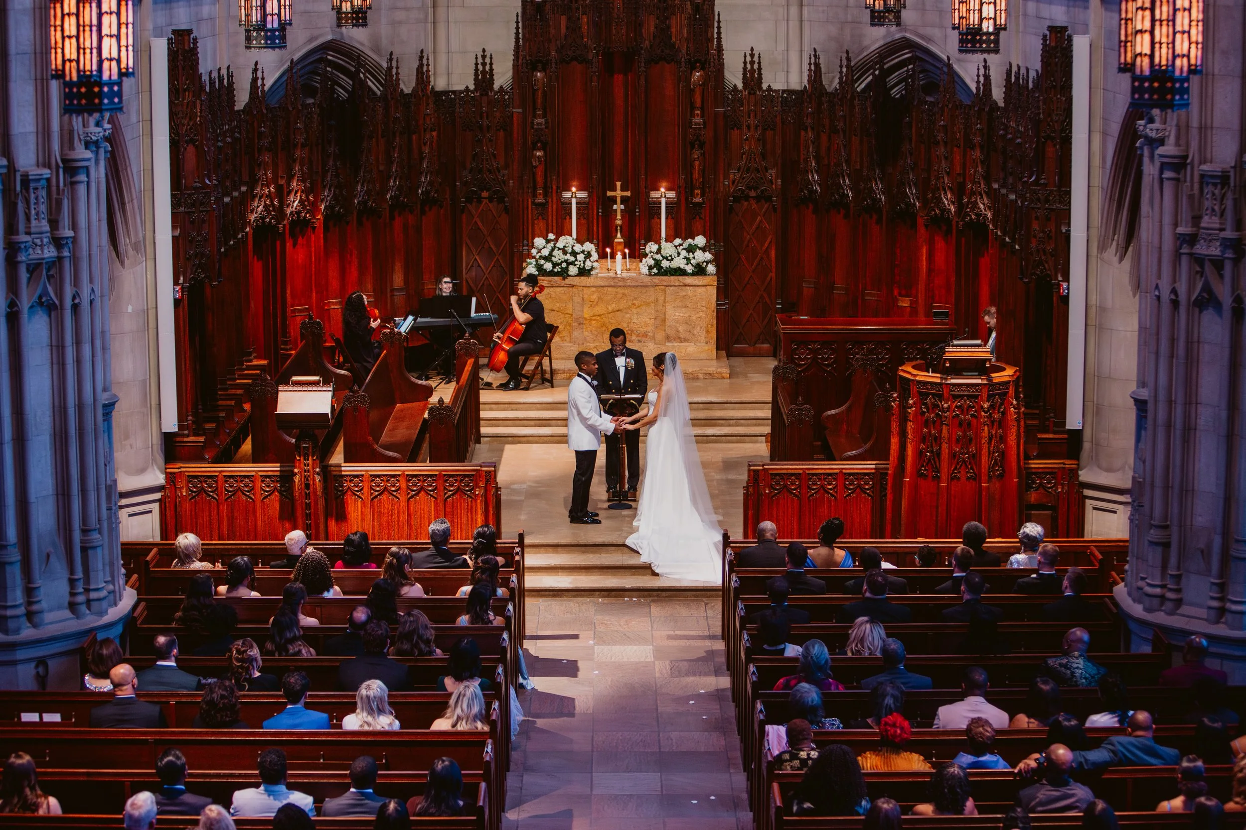 A wedding ceremony taking place inside Heinz Chapel. The bride and groom hold hands while Radiant Strings plays to the left of the altar.