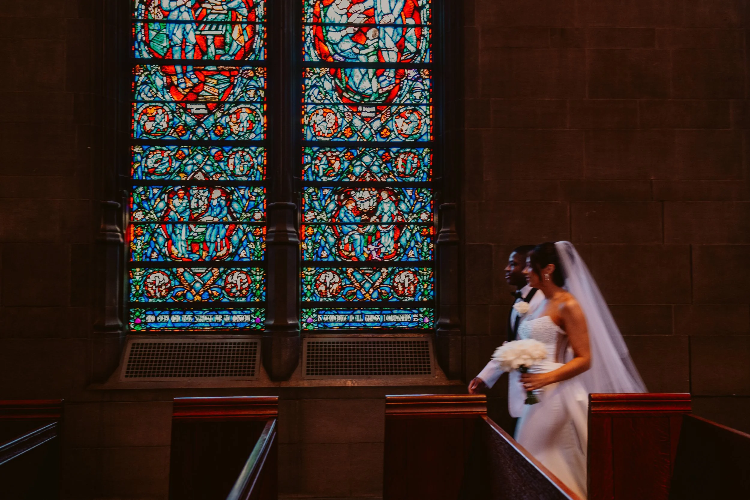 Melina and Drew walking through Heinz Chapel in their wedding attire next to a large, stained glass window.
