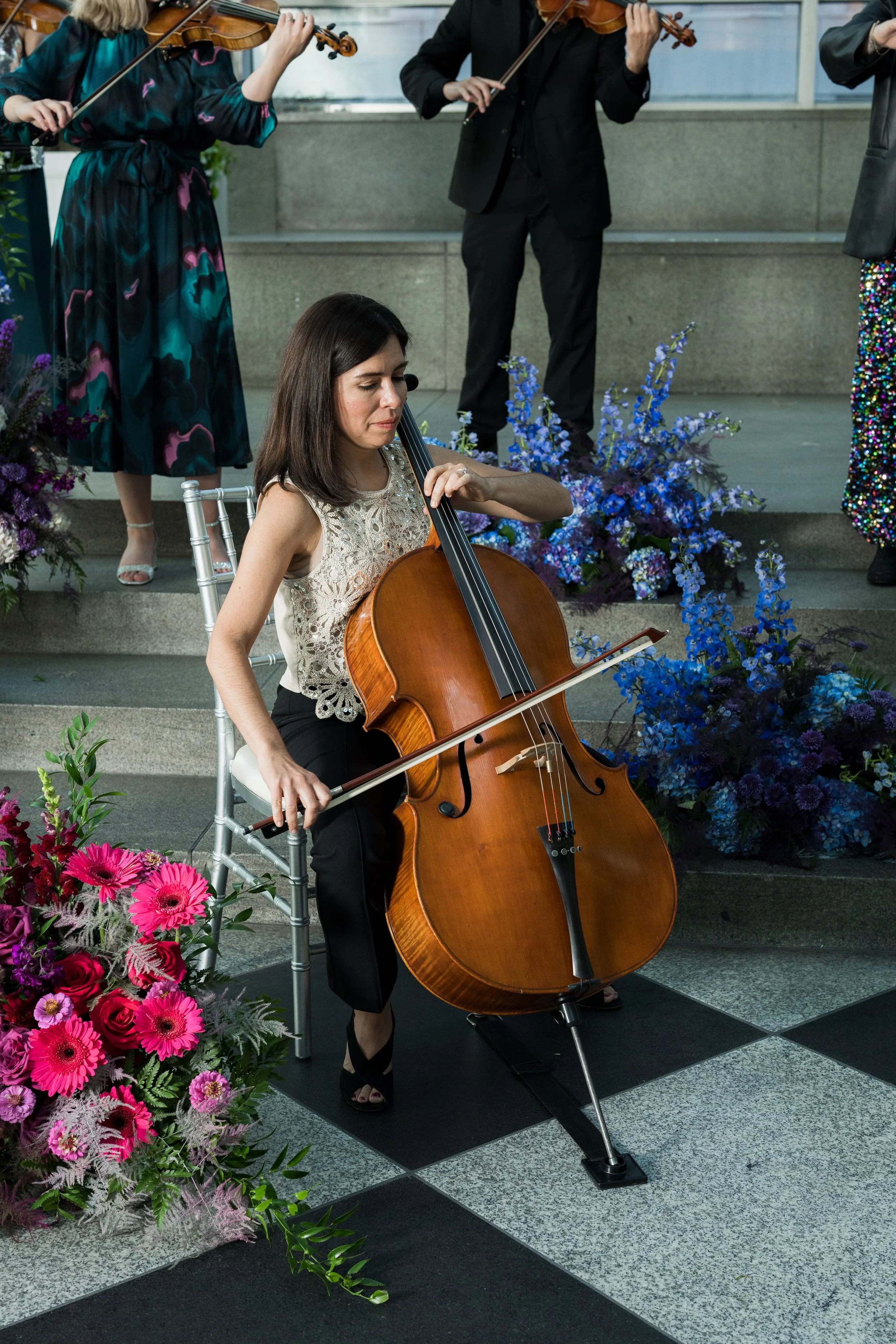 Becky Keeley playing a cello, sitting on a silver chair, surrounded by colorful flowers, at an indoor event with other musicians playing violins in the background.