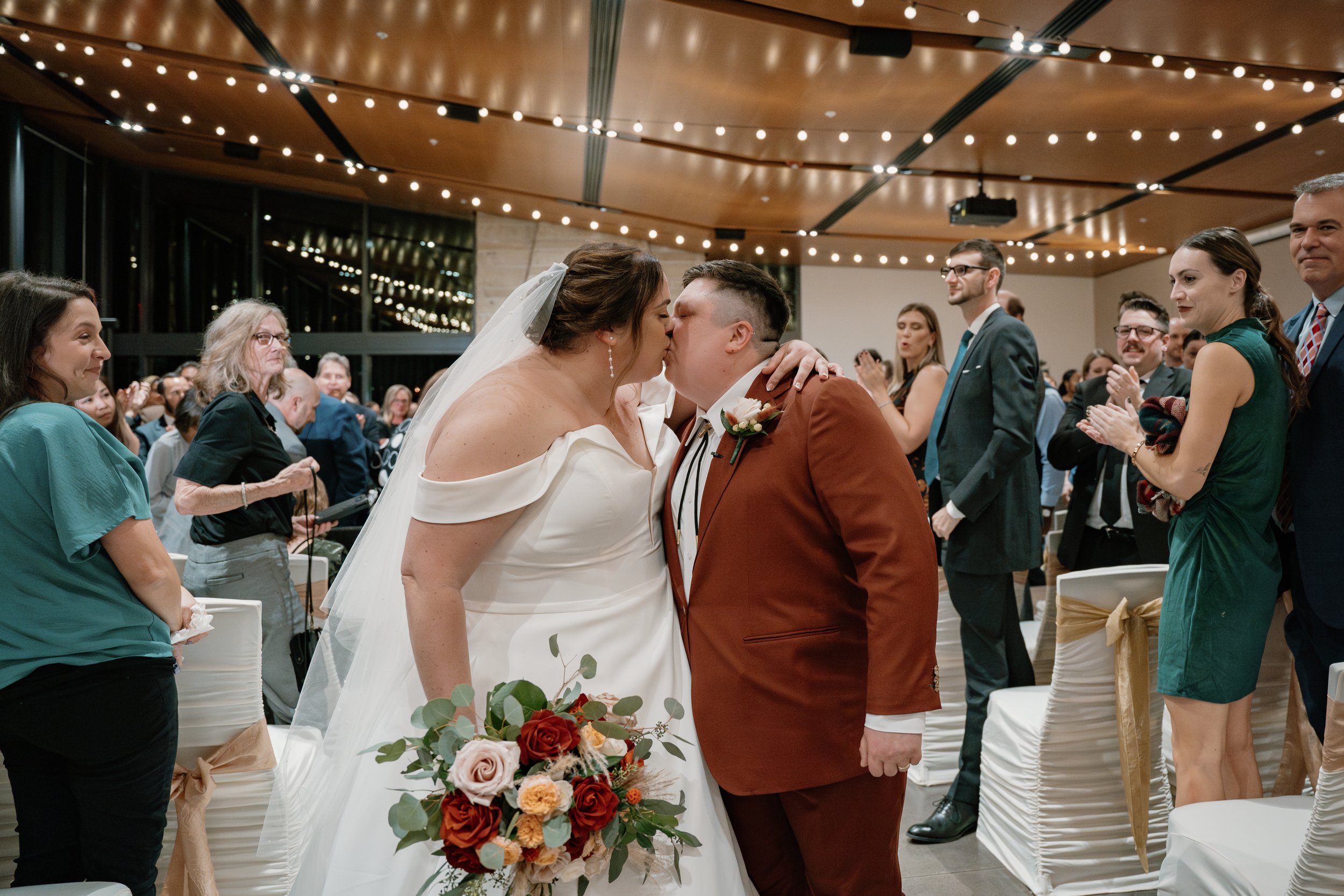 A bride and groom kiss during their wedding ceremony, surrounded by seated guests in an elegantly decorated venue with string lights overhead.