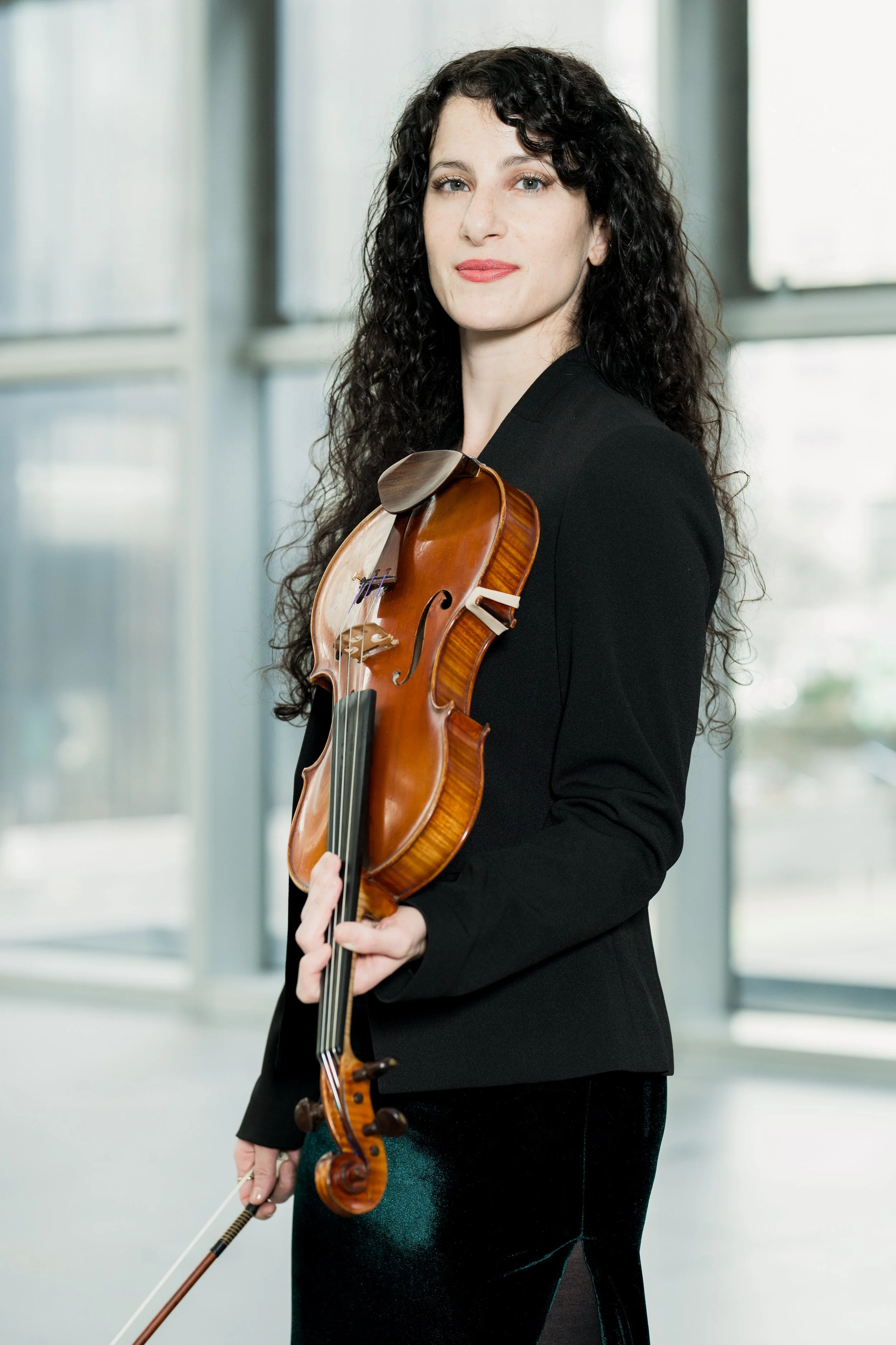 Cara Garofalo holding a violin, standing indoors with large windows behind her.