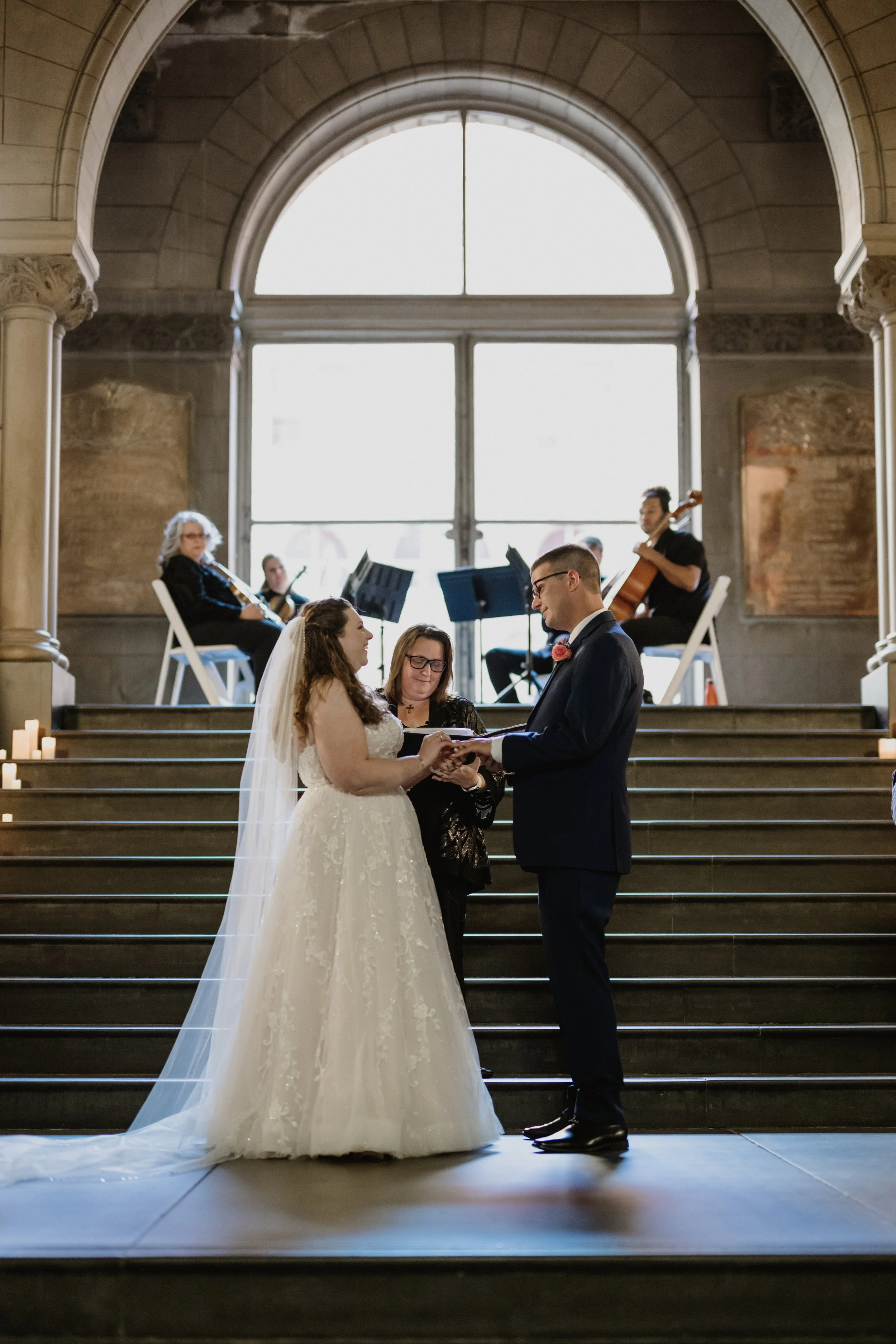 Nicole and Devin exchanging vows during their wedding ceremony on stairs of the Allegheny County Courthouse, with Radiant Strings playing string instruments in the background.