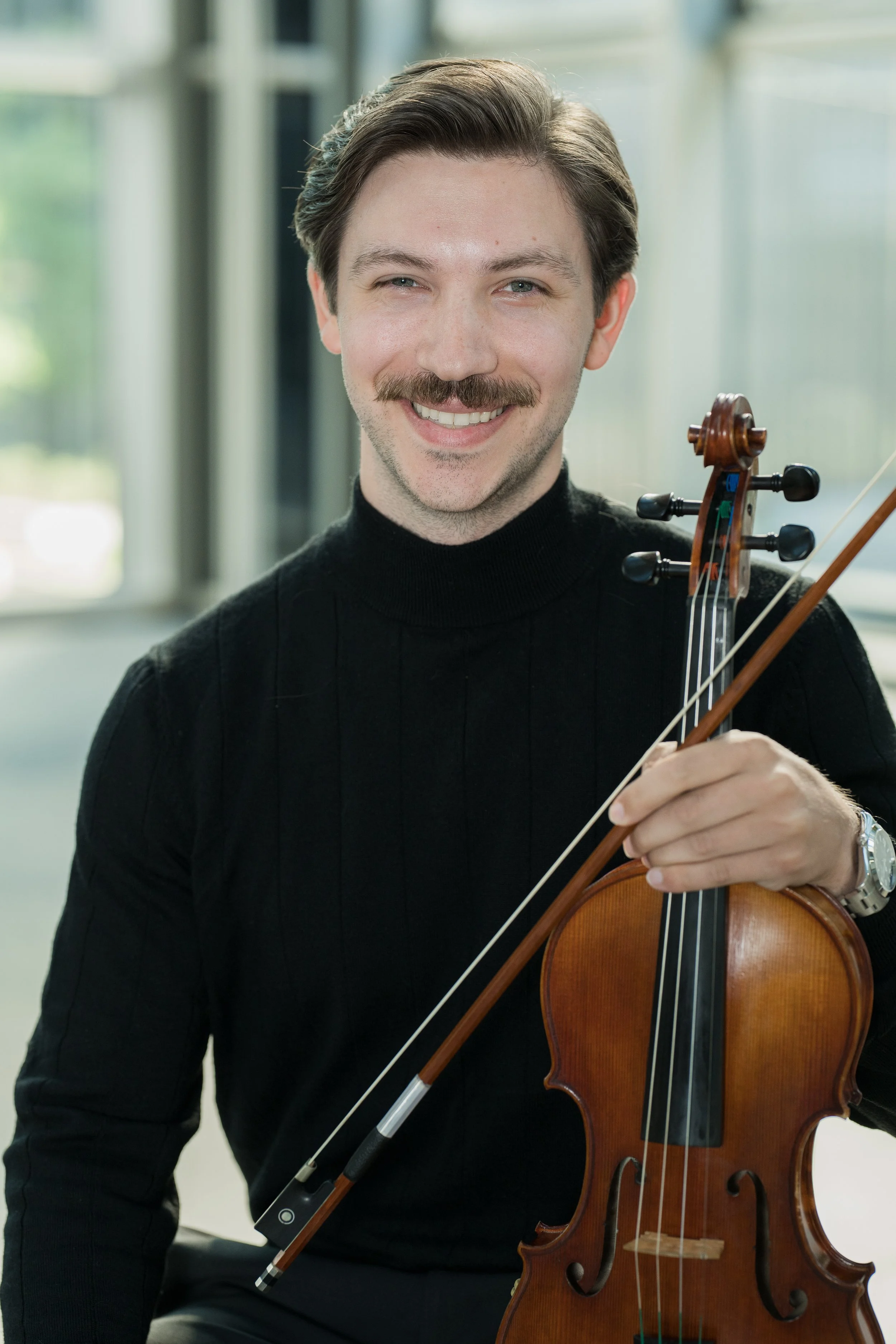 Anthony Cicco smiling, holding a violin and a bow, dressed in a black turtleneck, standing indoors in front of a window.