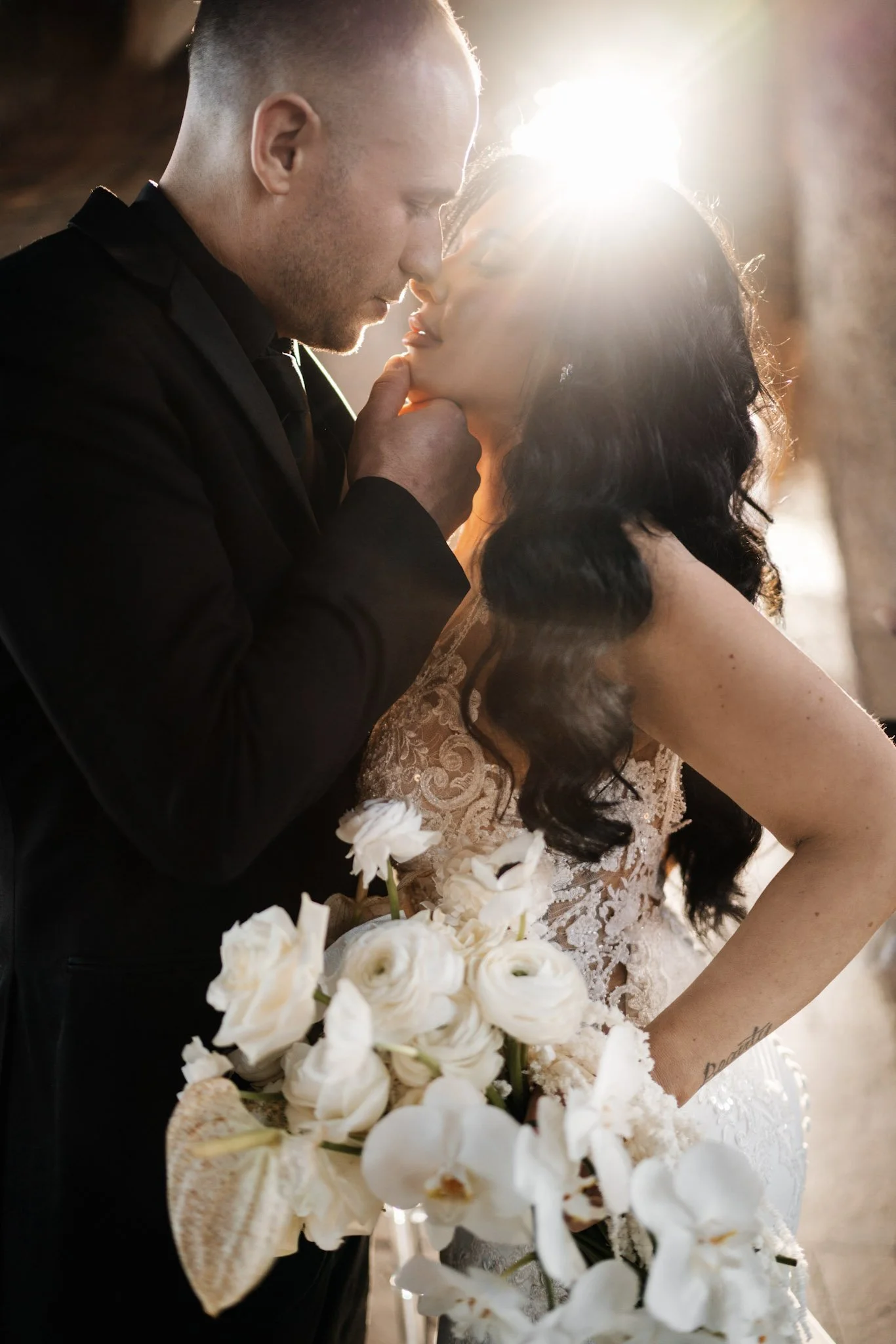 Amanda and Cory about to kiss. Amanda is holding a bouquet of white flowers and wearing a lace wedding dress. Bright sunlight is shining behind them, creating a romantic glow.