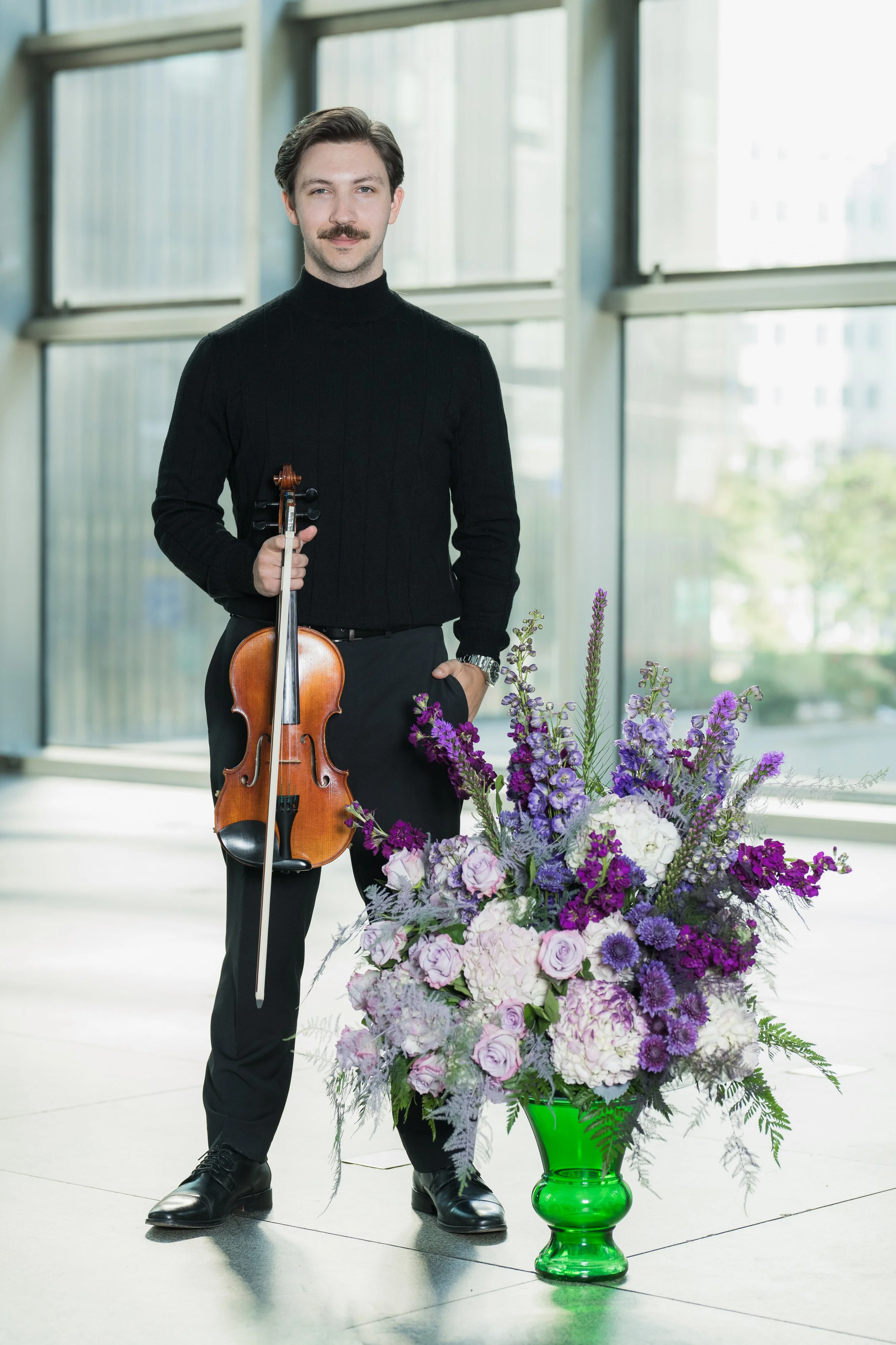 Anthony Cicco holding a violin stands next to a large bouquet of purple and white flowers in a green vase, inside a modern building with large windows.