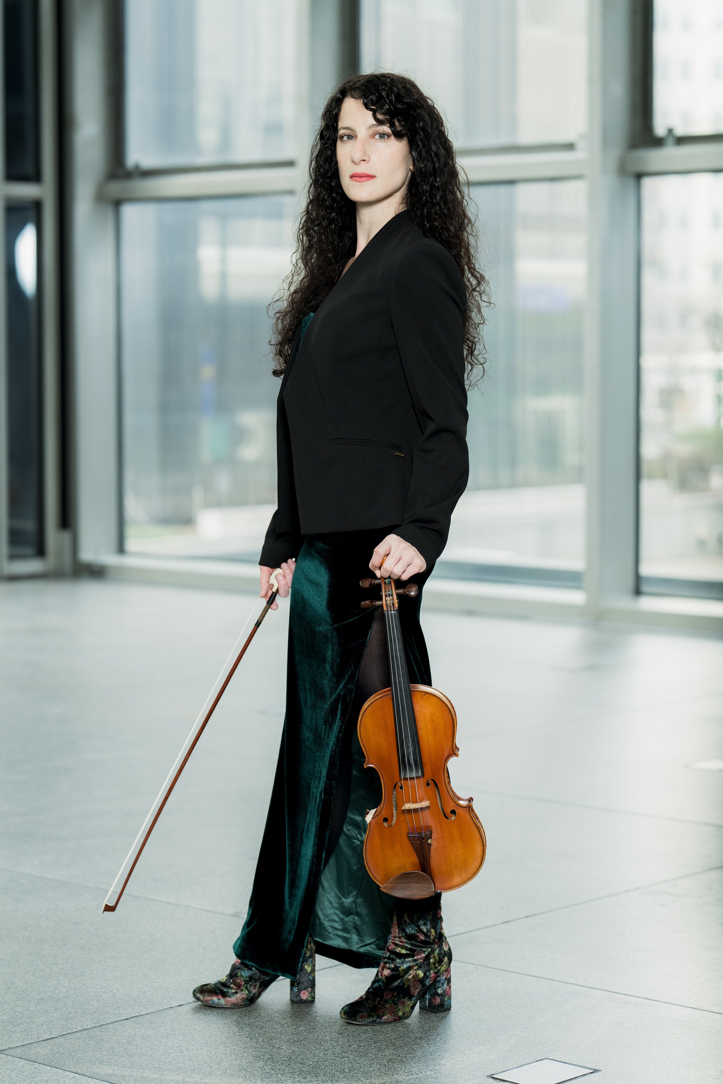 Cara Garofalo holding a violin and bow, standing in a modern indoor space with large windows.