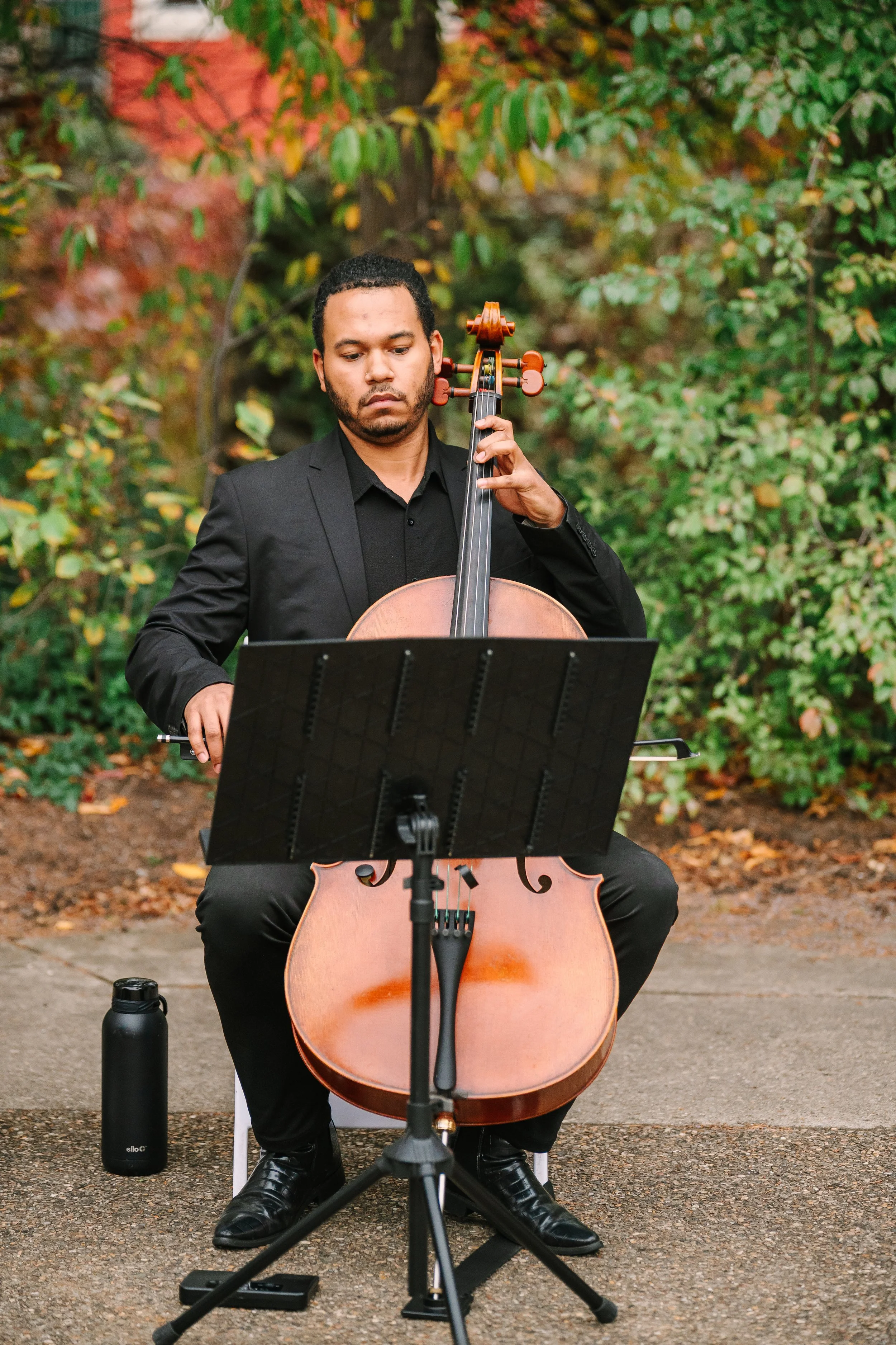 Ahmer'e Blackman playing a cello outdoors, surrounded by autumn-colored foliage.