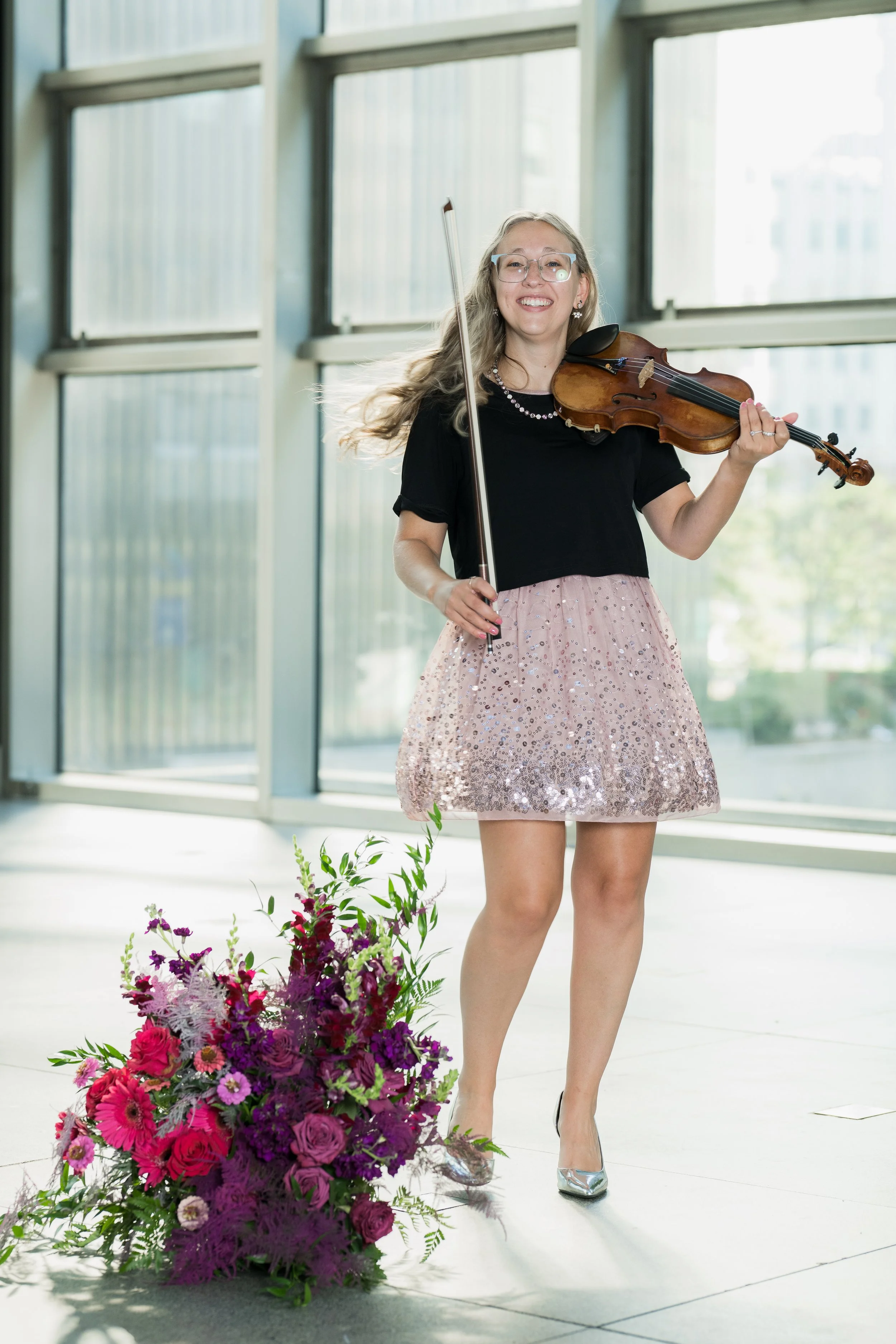 Samantha Smith holding a violin and bow, standing next to a large bouquet of pink and purple flowers inside a modern building with large glass windows.