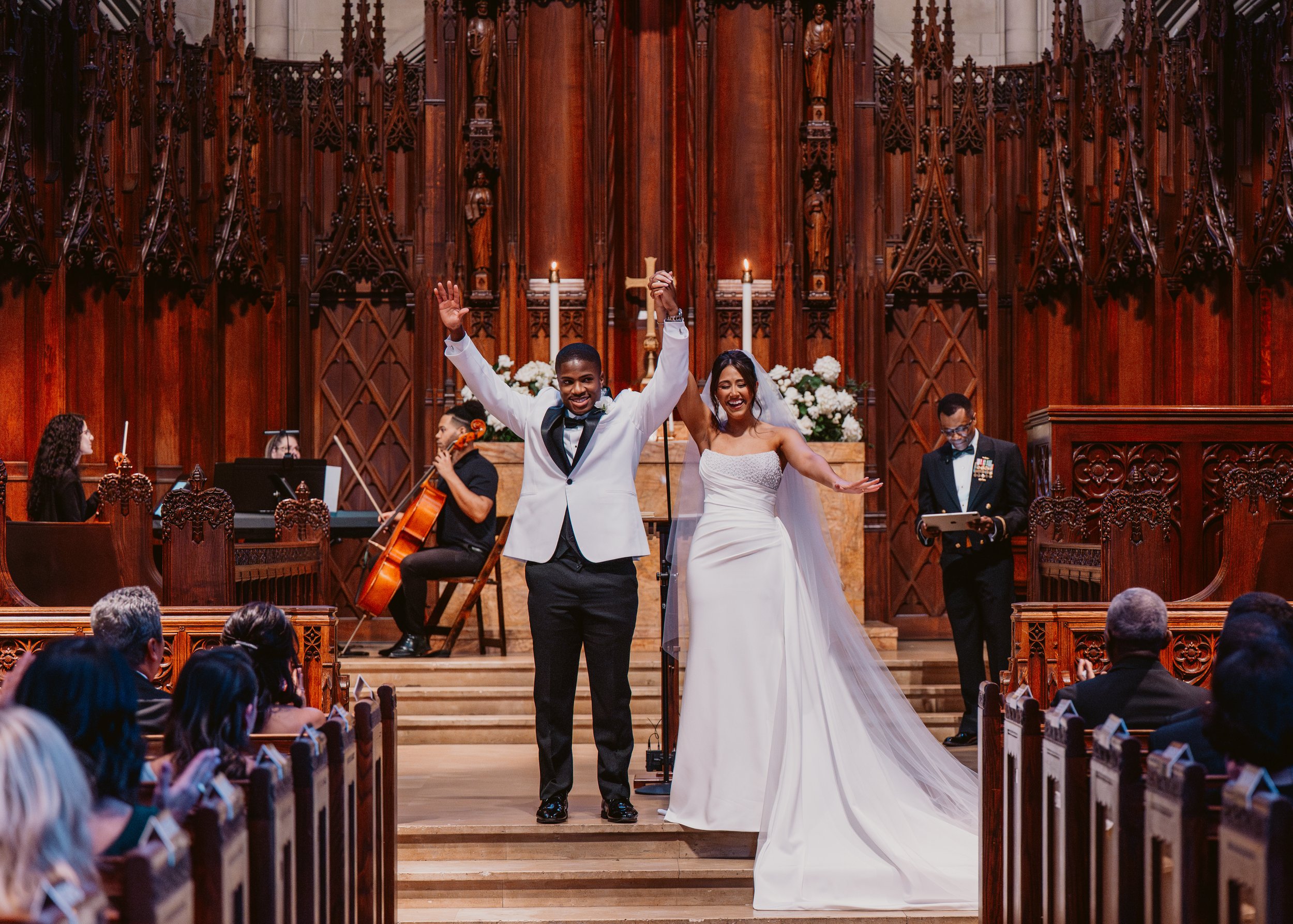 A newlywed couple celebrates their wedding victory in a church with a wooden interior. The groom in a white tuxedo jacket and black pants, and the bride in a white strapless wedding gown, stand hand-in-hand at the altar, smiling. Musicians, including a cellist and pianist, are seated behind them. Guests seated in pews watch and take photos.