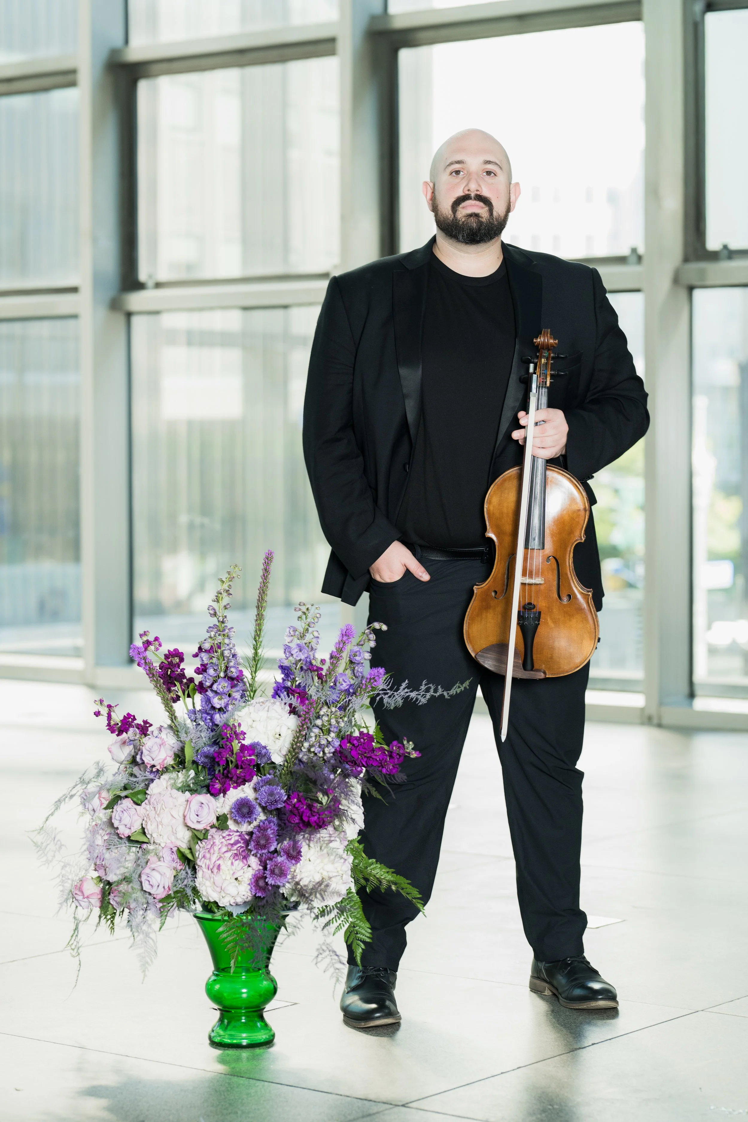Stephen Weiss holds his violin near a large bouquet of purple and white flowers in a green vase.