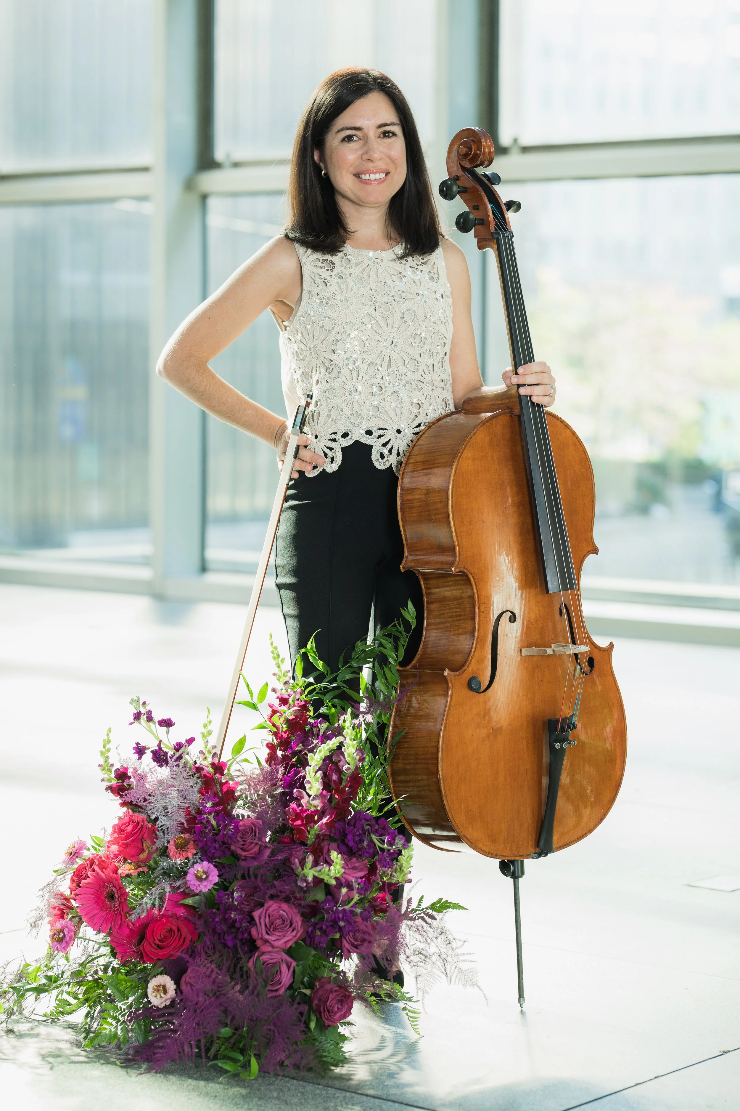 Becky Keeley holding a cello, standing next to a large bouquet of pink and purple flowers, inside a modern building with large windows in the background.