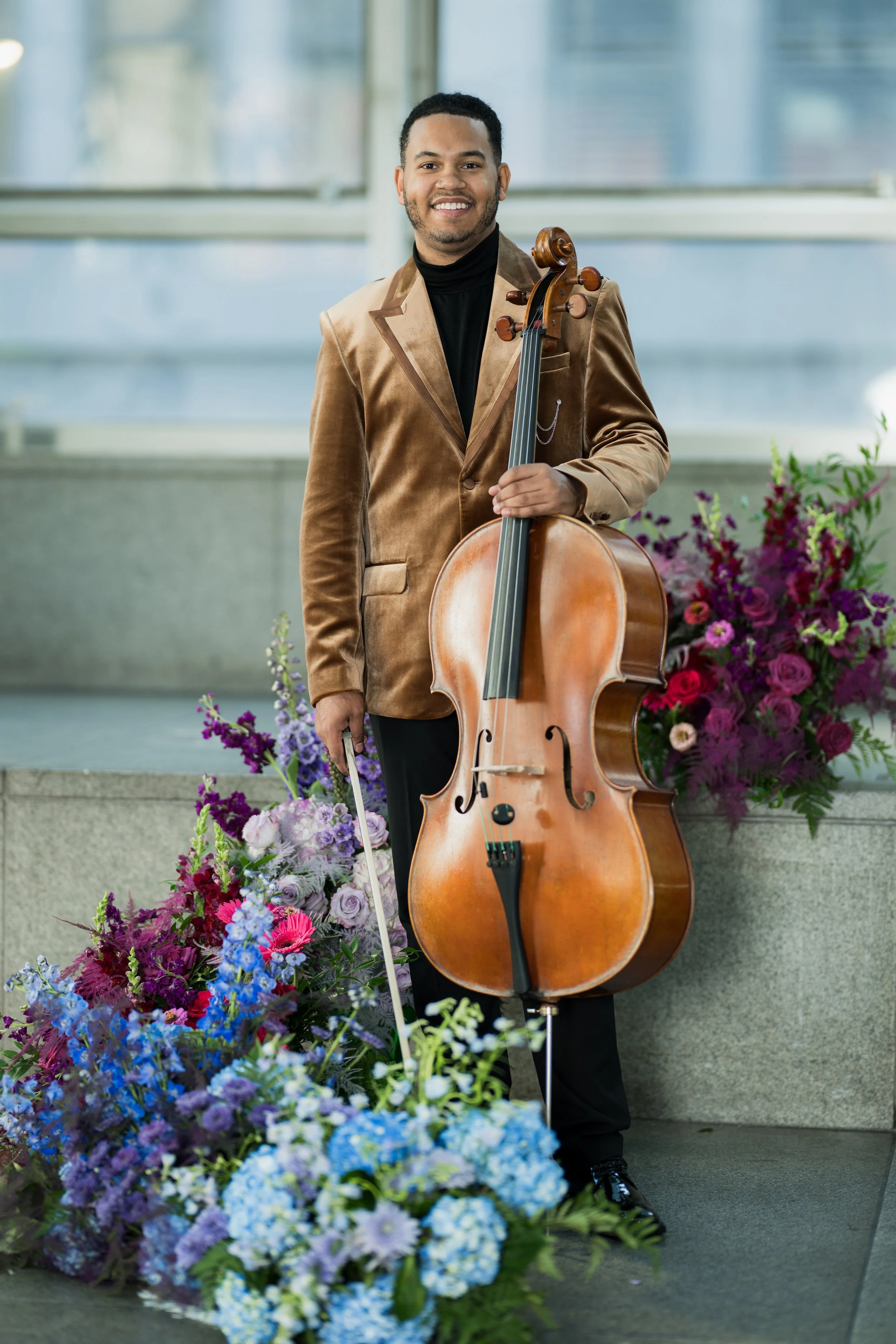 Ahmer'e Blackman holding a cello and bow, standing in front of a large arrangement of purple, pink, and blue flowers indoors.