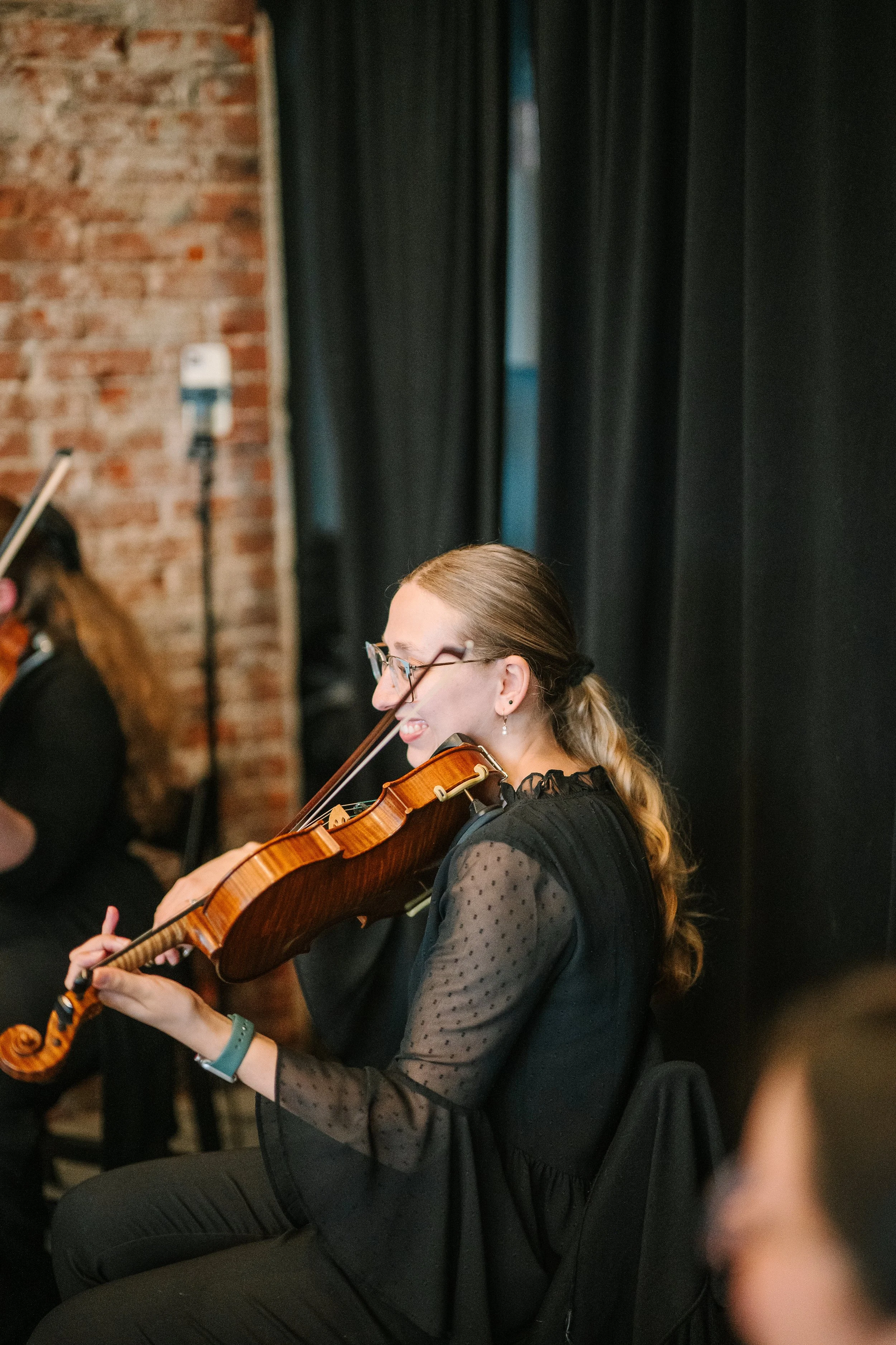 Samantha Smith of Radiant Strings playing a violin during a performance, sitting on a chair in a room with black curtains and a brick wall.