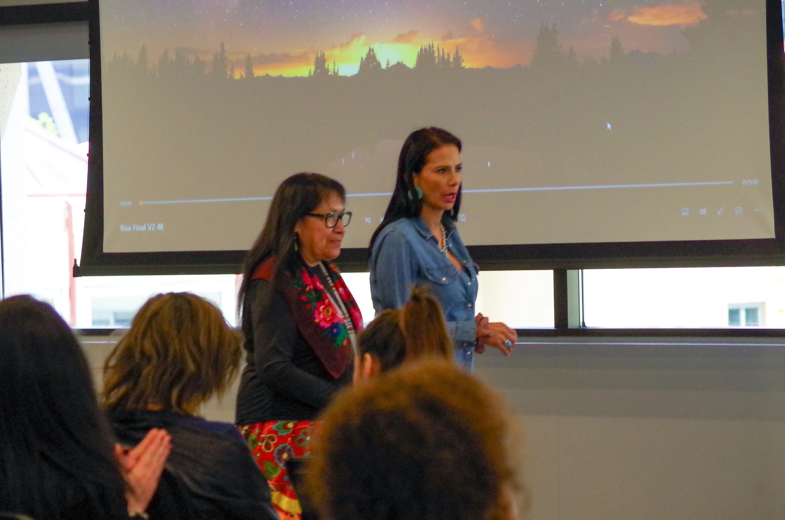 Two women standing in front of a large screen in a conference room, addressing an audience. The screen displays a sunset landscape with trees.