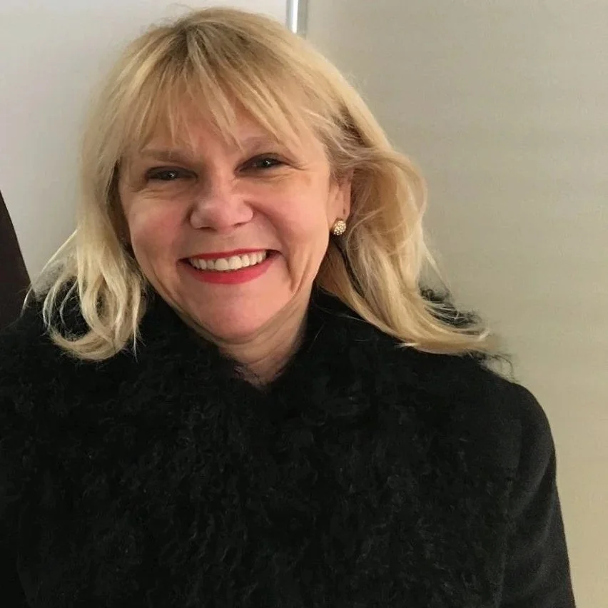 A woman with blonde hair smiling at the camera, wearing a black top and earrings, standing against a neutral background.