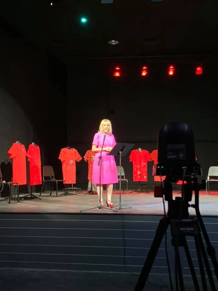 A woman in a pink dress standing at a microphone on a stage, with red dresses displayed on mannequins behind her. There is a camera in the foreground focused on her.