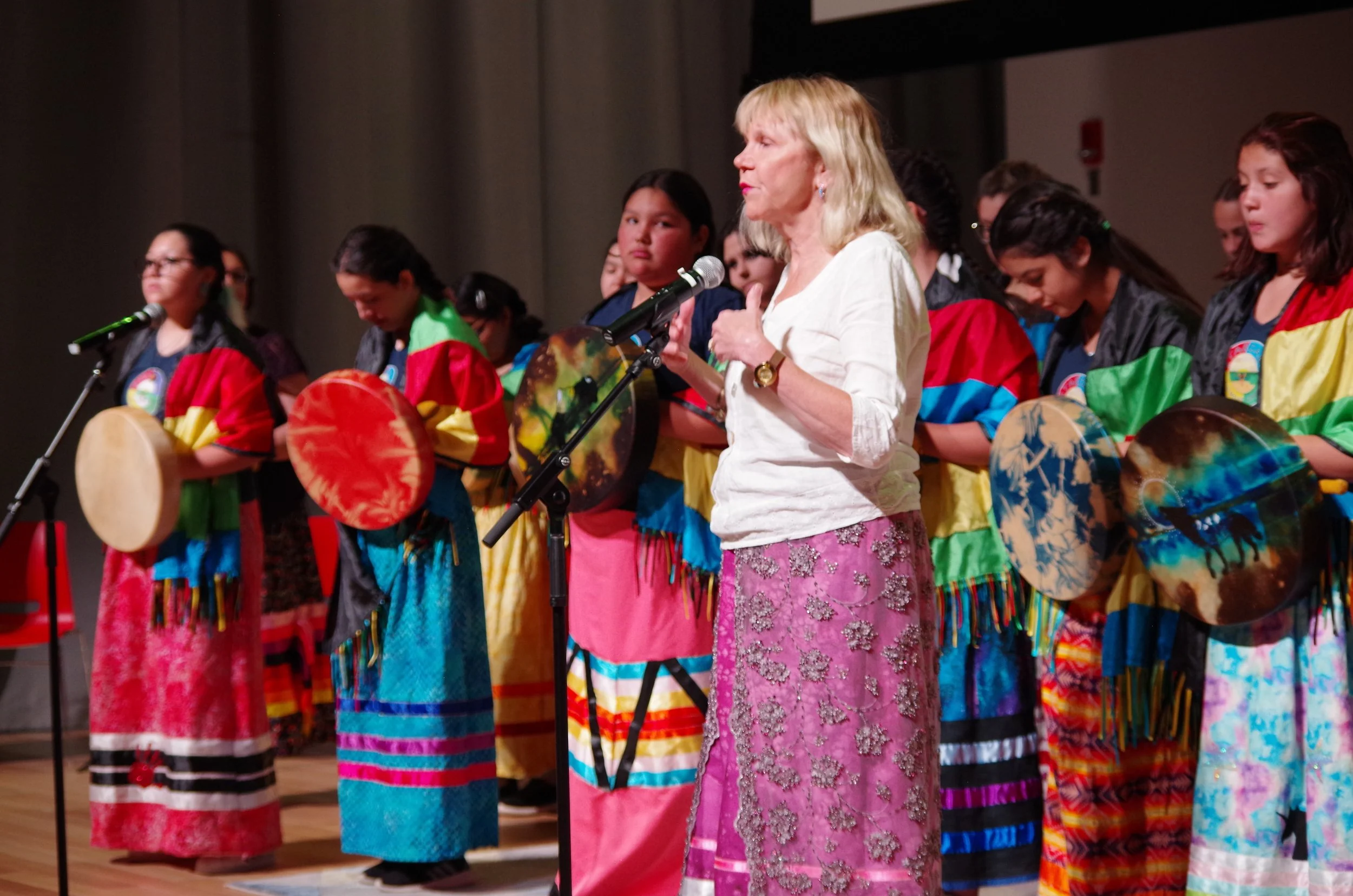 A group of young girls in colorful traditional clothing holding drums, standing on stage during a performance, with an older woman speaking into a microphone.