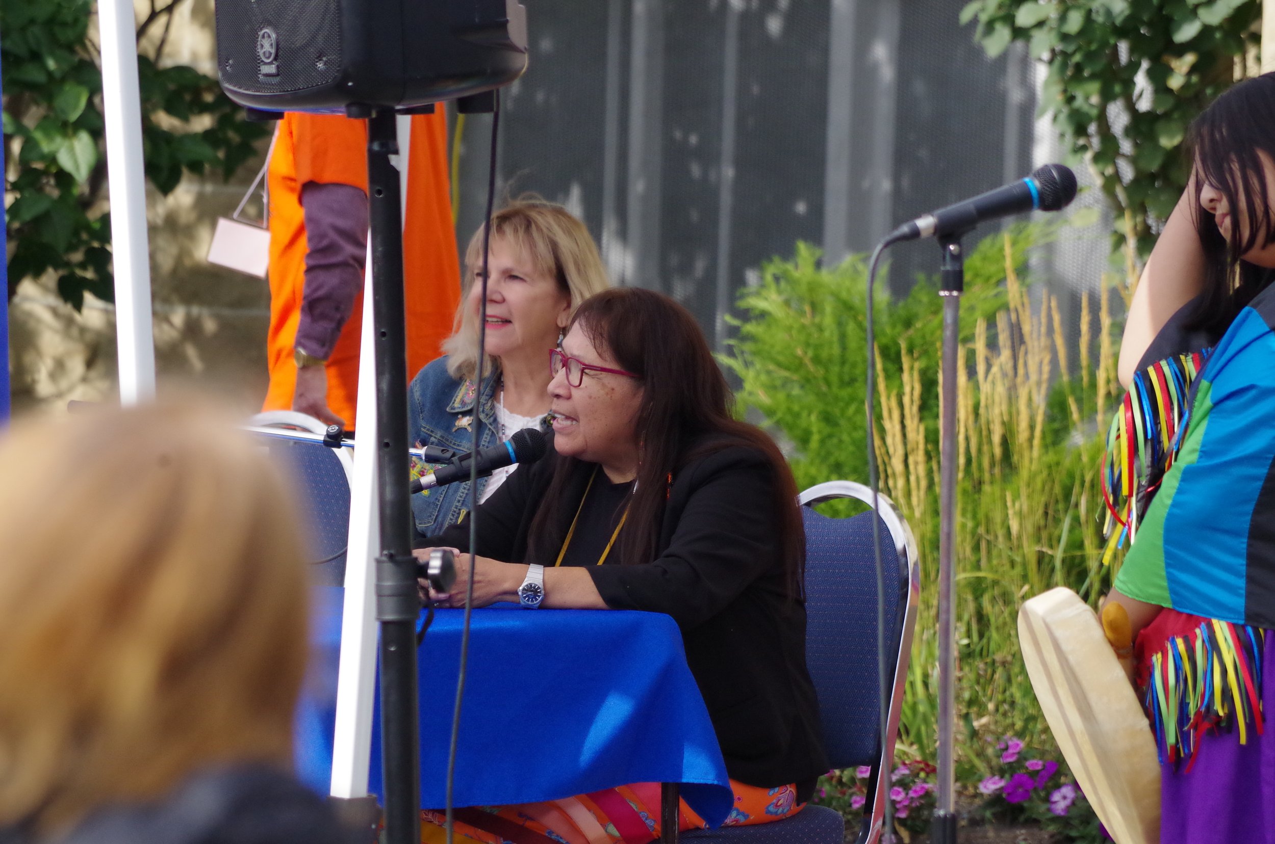People speaking at an outdoor event, with microphones, a blue tablecloth, and greenery in the background.