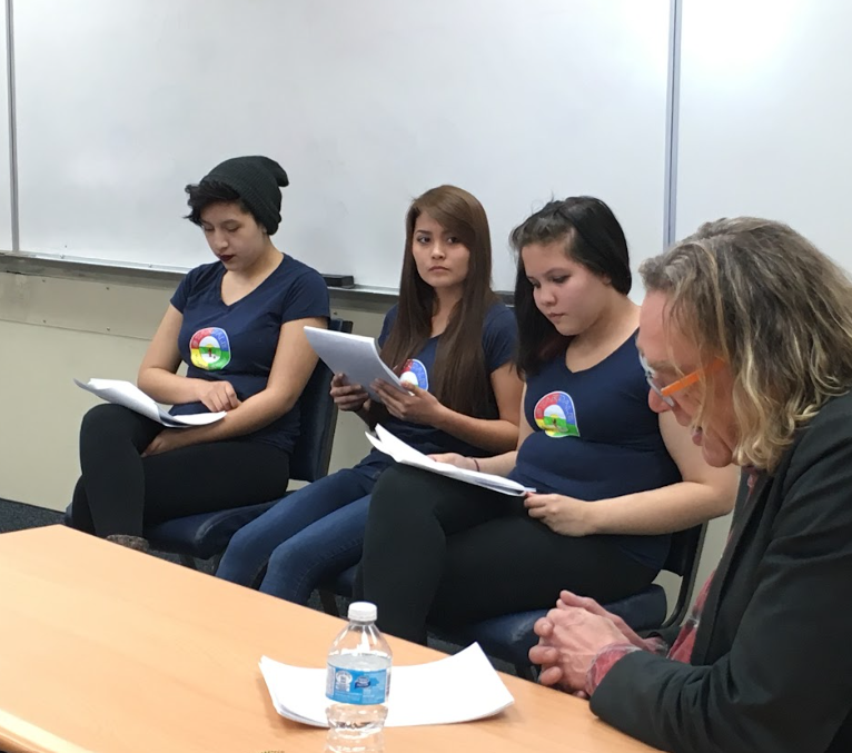 Group of four young women sitting on chairs, reading papers, with a woman at the end of the row in a business suit and glasses, in a classroom or meeting room setting.