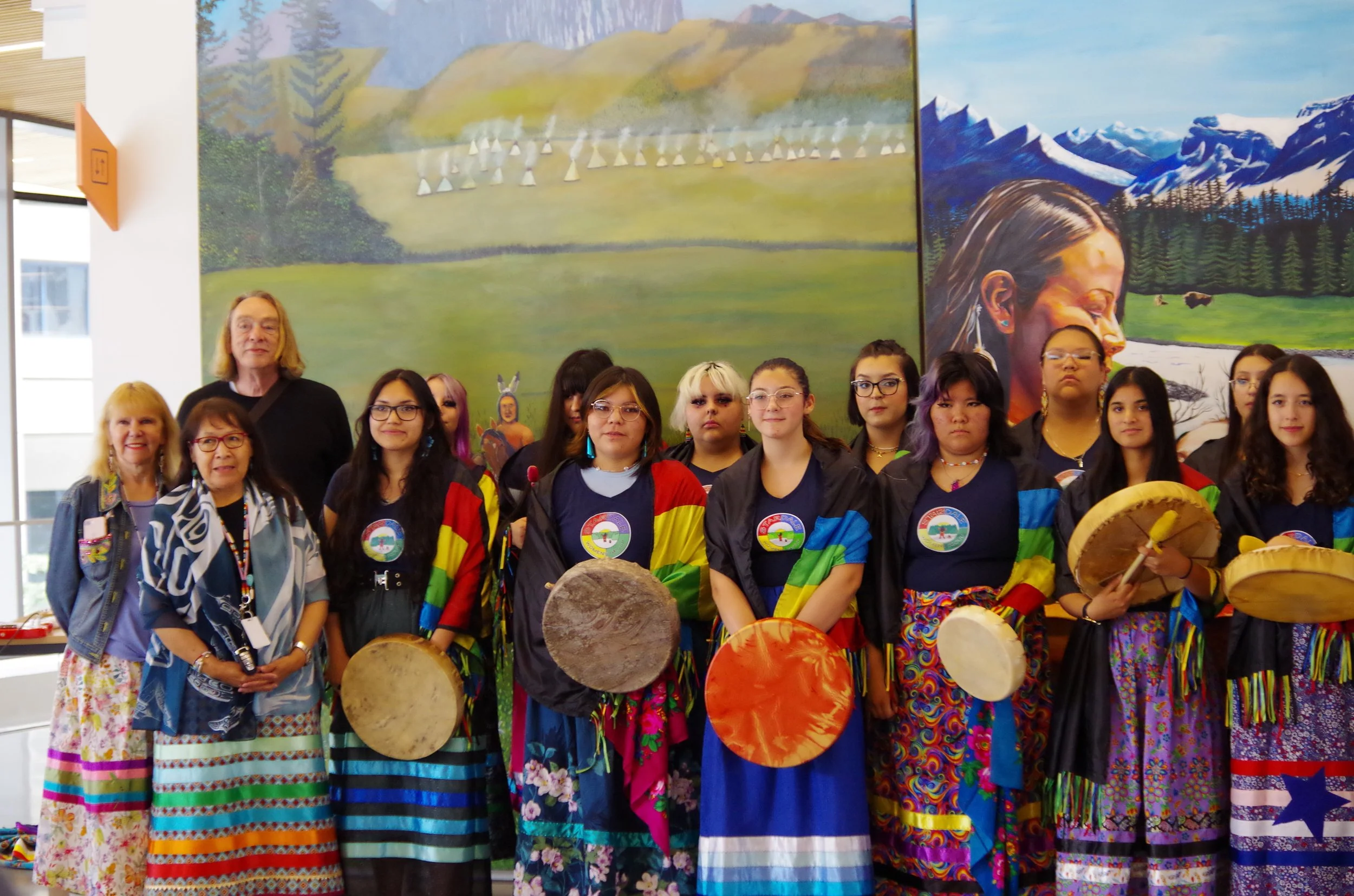 Group of women and young girls wearing colorful traditional and modern clothing, some holding drums, standing in front of a mural painting featuring a landscape of mountains, trees, and a indigenous woman with a mountain landscape in the background.