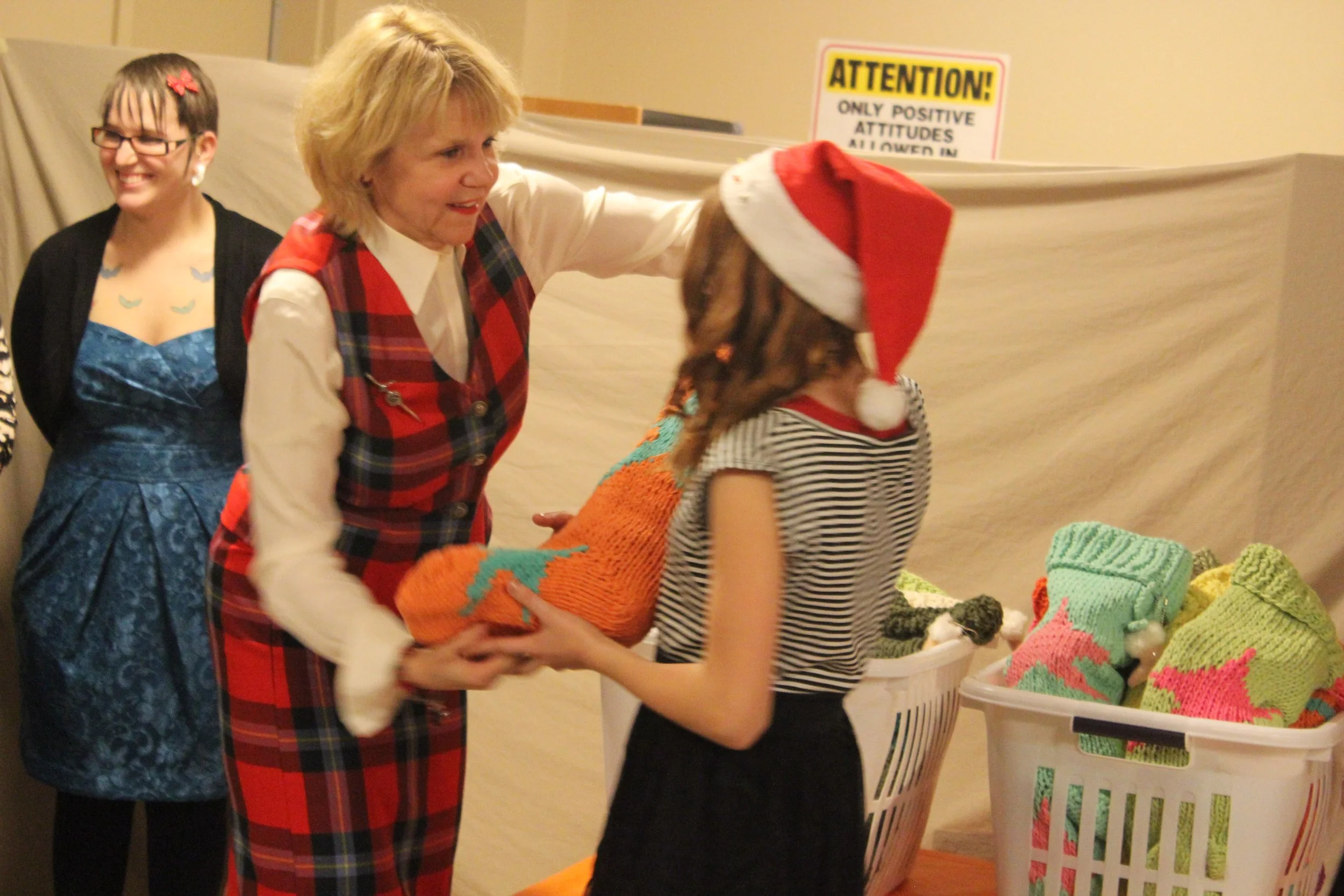 A woman in a red plaid vest giving a colorful Christmas stocking to a girl wearing a Santa hat and striped shirt. A woman in a blue dress and glasses is smiling in the background. There is a basket of knitted Christmas stockings nearby.
