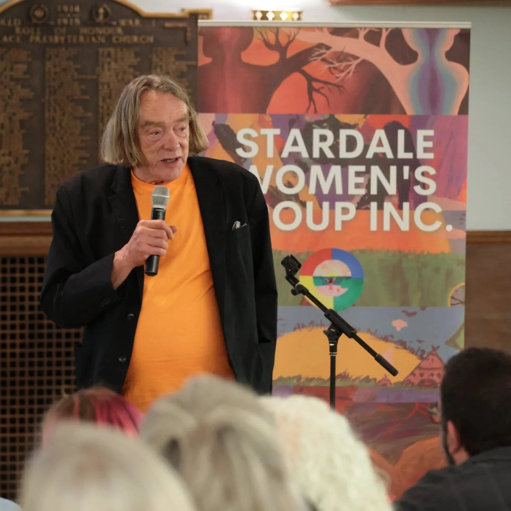 A man with shoulder-length gray hair is speaking into a microphone at an event for Stardale Women's Group Inc. behind him is a colorful banner with the group's name and a painting of a tree and landscape.