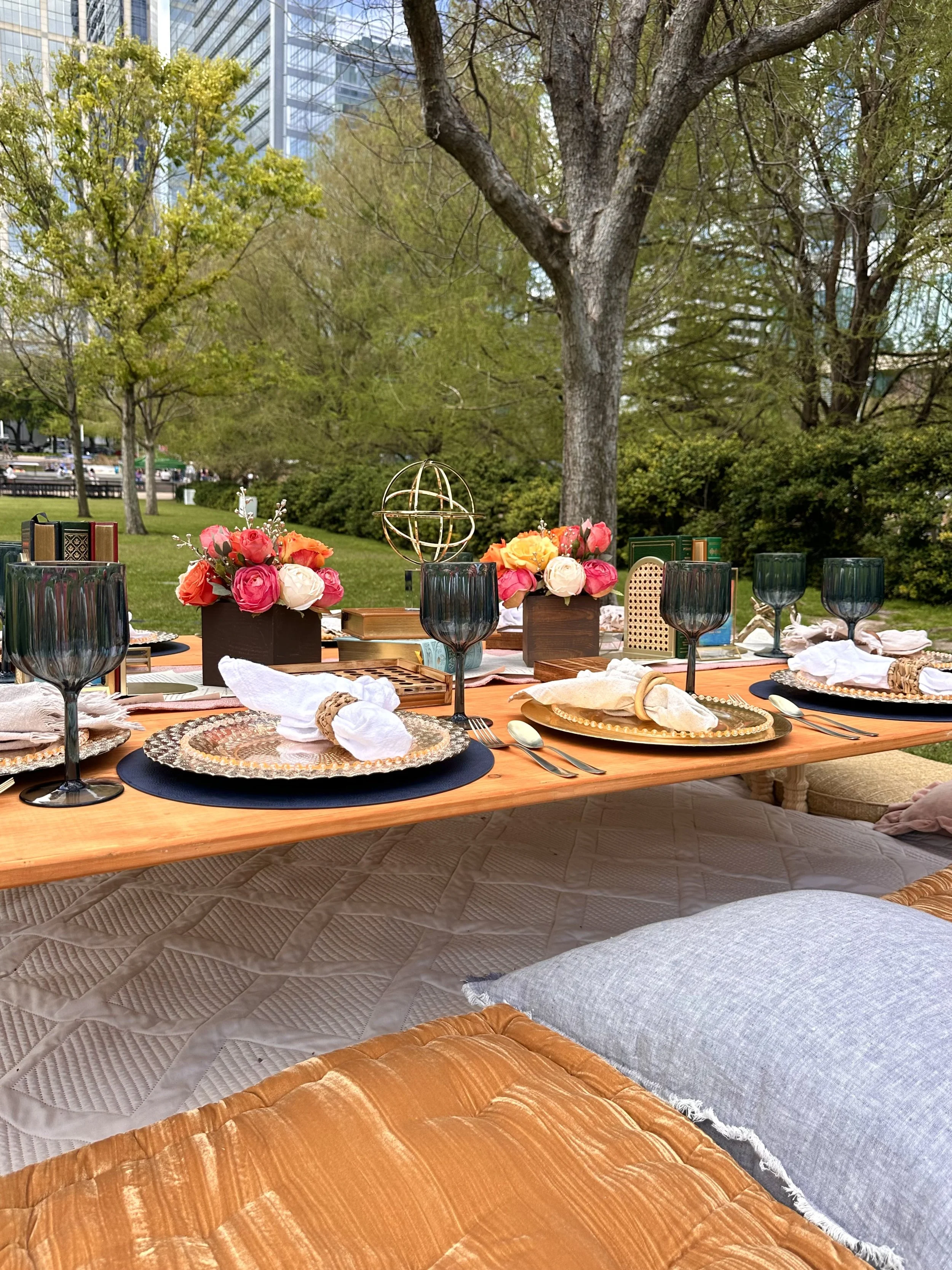 A daytime outdoor event setup with a table of floral decorations, candles, and tableware on a wooden table in a park with trees and tall buildings in the background.