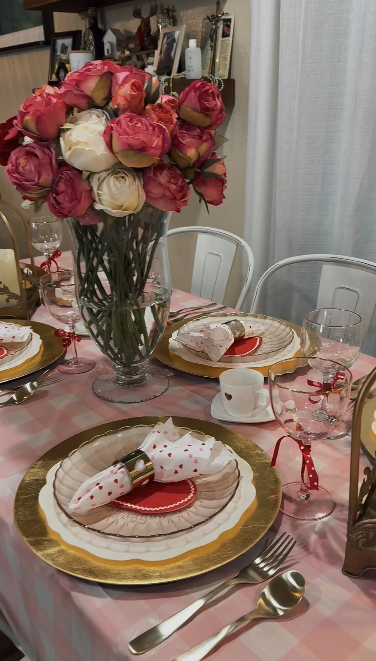 A decorated dining table with a pink and gold theme, featuring a large bouquet of pink and cream roses in a tall glass vase. The table has gold-rimmed plates, pink and clear glasses with red ribbons, white napkins with red hearts, and gold flatware. 