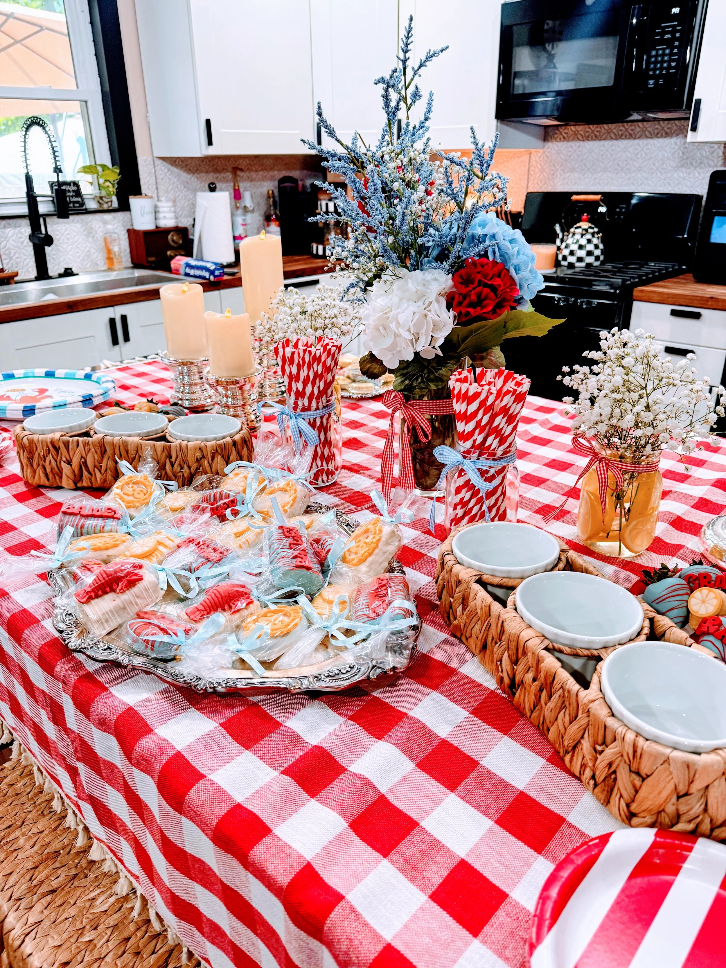 A kitchen table with a red and white checkered tablecloth, decorated for a celebration with flowers, candles, and baked goods, set in a bright kitchen with white cabinets and black appliances.