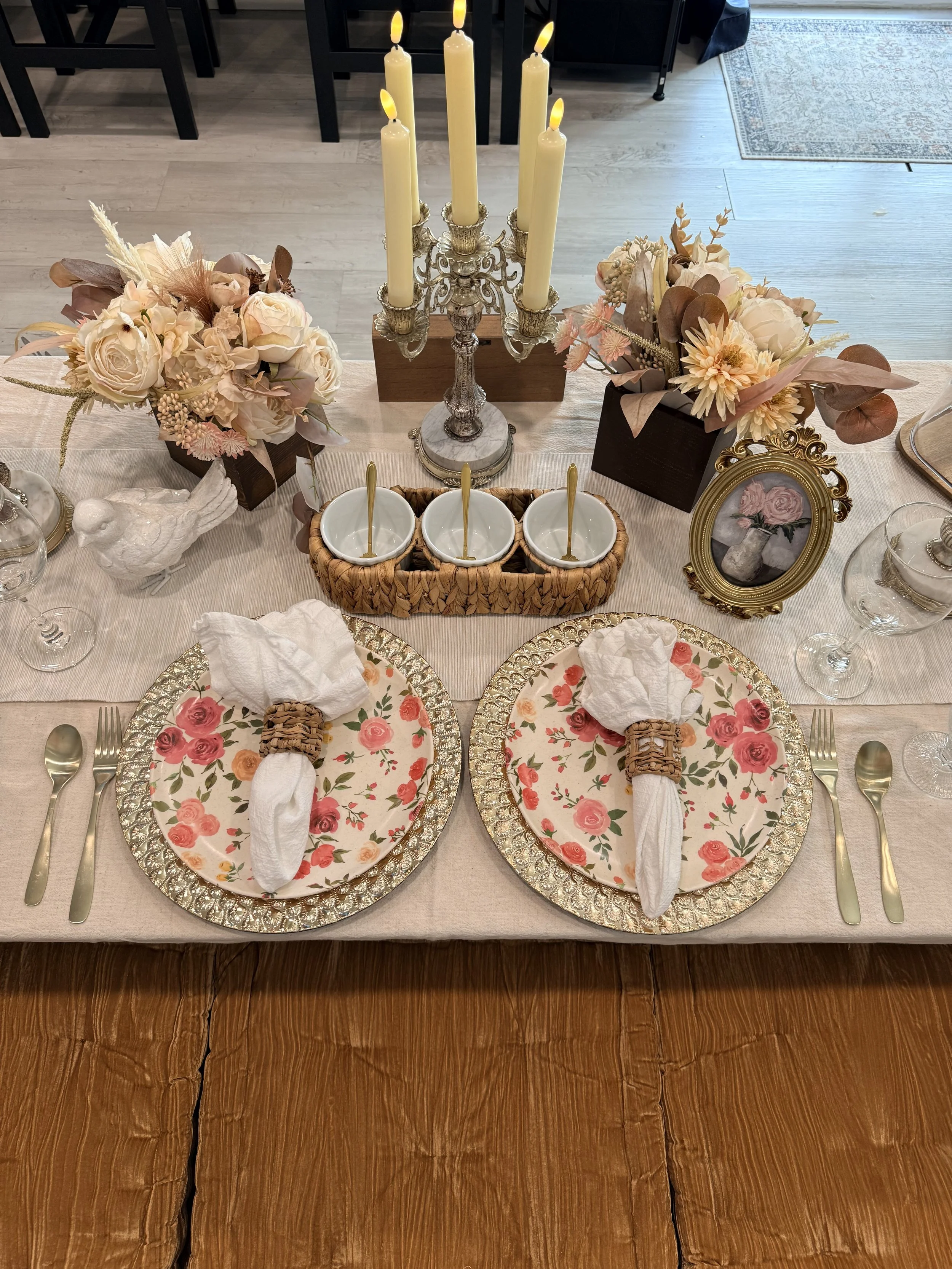 A dinner table with floral-themed plates, white napkins in basket holders, gold cutlery, and decorative items including a white bird sculpture, floral arrangements, a candelabra with lit candles, and a small framed painting of a vase with roses.