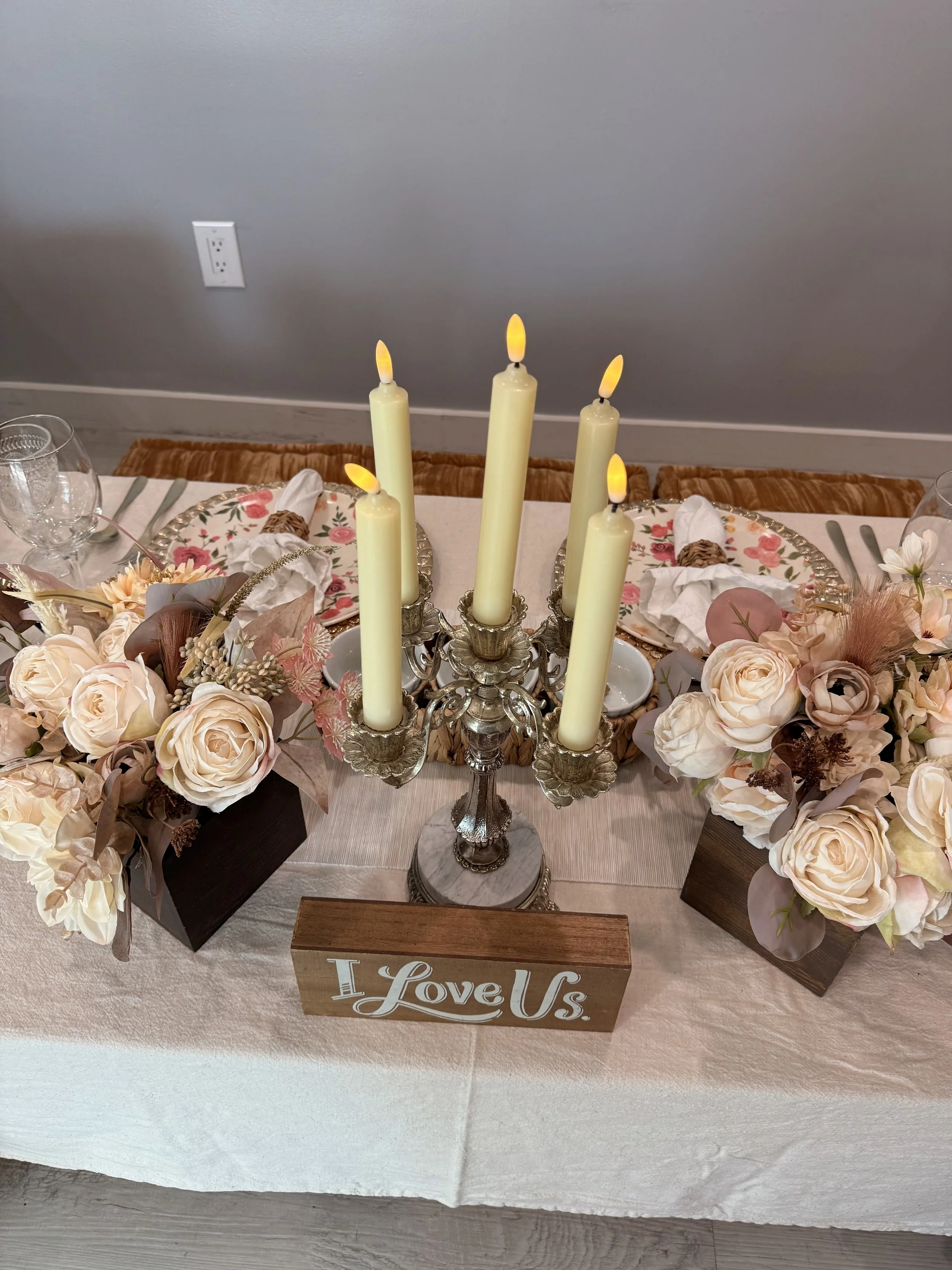 A decorative table centerpiece with a silver candelabrum holding five LED candles with flickering flames, surrounded by floral arrangements of pale pink and cream roses and other flowers, on a table with a cream tablecloth. There is a wooden sign wit