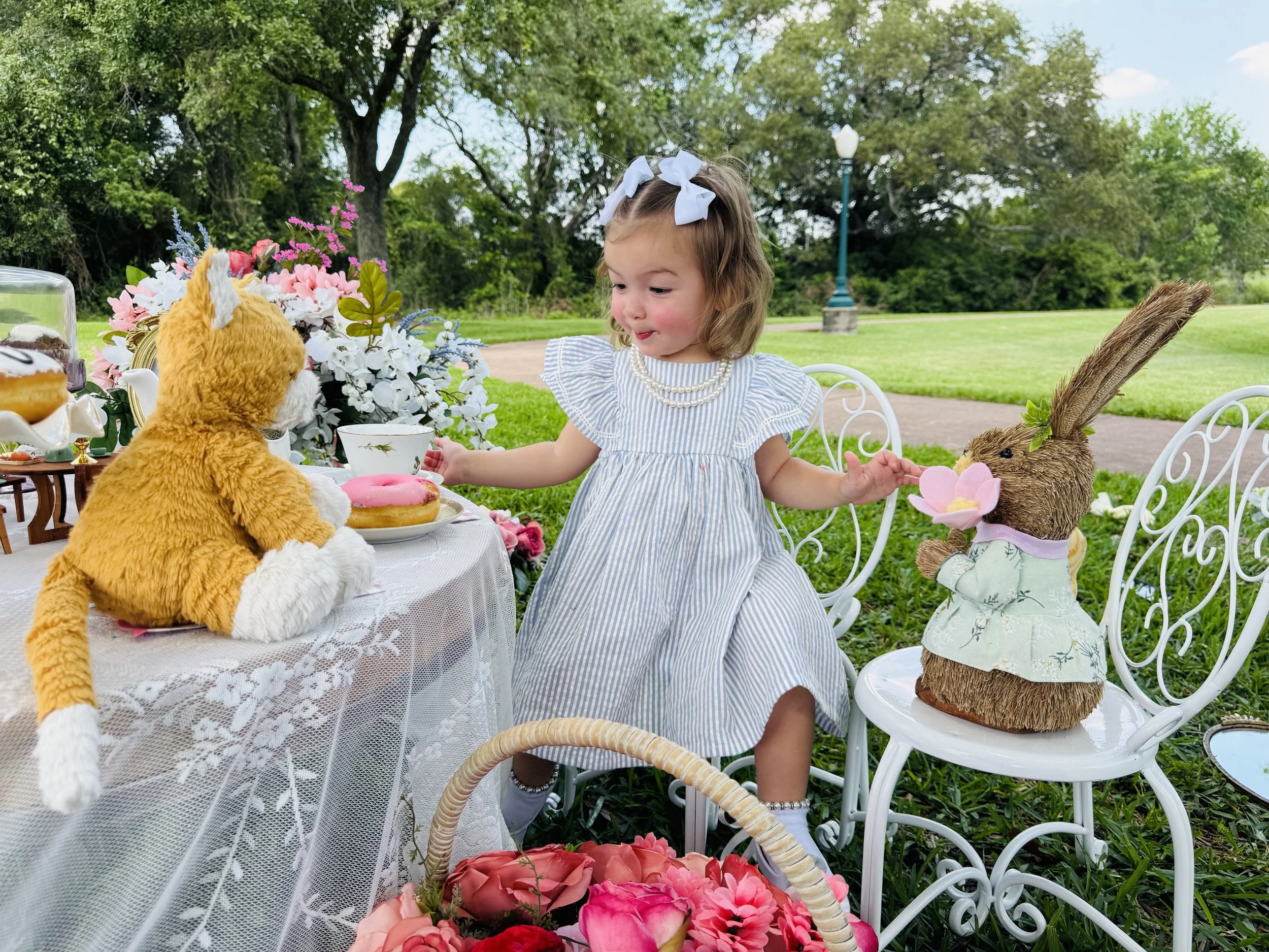 A young girl with curly hair, wearing a striped dress, pearl necklace, and white bow in her hair, sitting at a lace-covered table outdoors with flower arrangements and plush bunny and tiger toys.