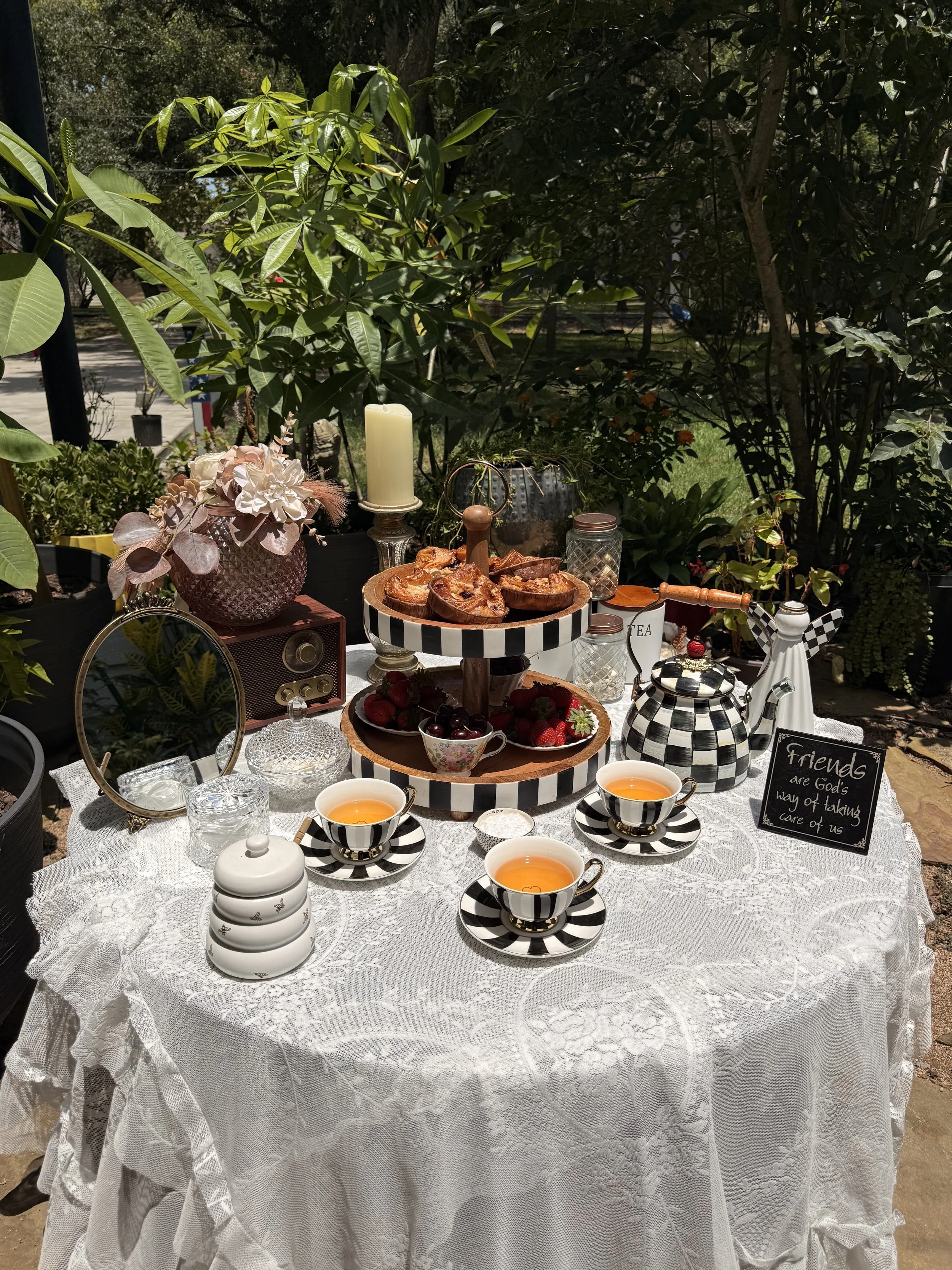 An outdoor table set with black and white striped teacups of tea, a white lace tablecloth, a white ceramic teapot, a white ceramic sugar container, a tiered stand with strawberries and cherries, a plate of assorted baked goods, a purple vase with a f