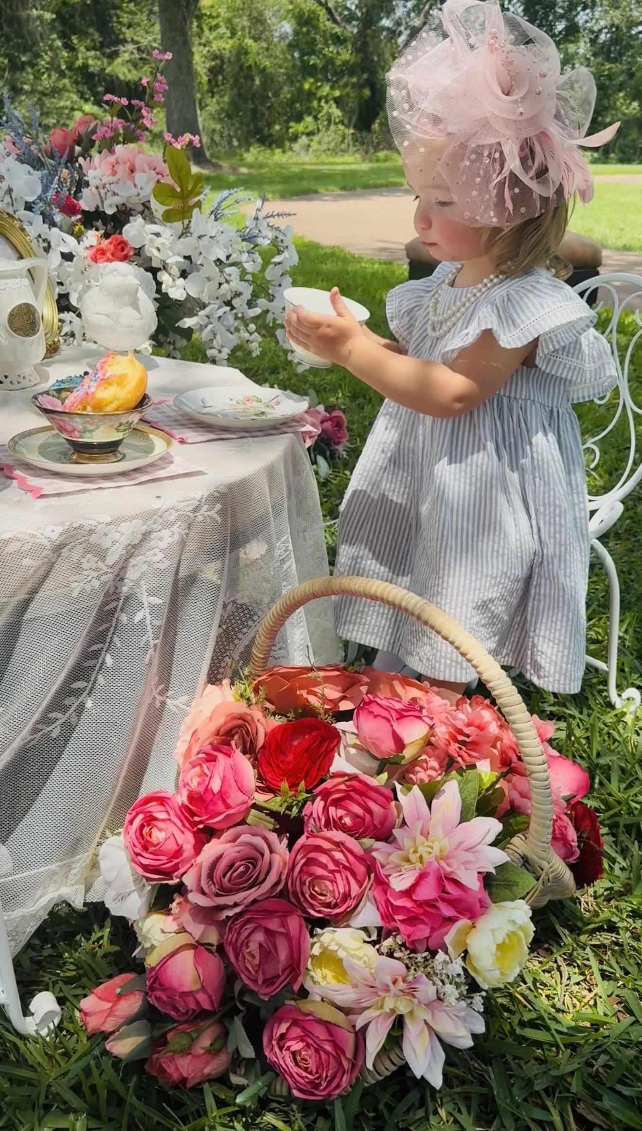 A young girl wearing a pink tulle hat, pearl necklace, and striped dress at an outdoor tea party. She is holding a teacup near a table decorated with flowers, cakes, and teapots. A basket of pink and red flowers is in the foreground.