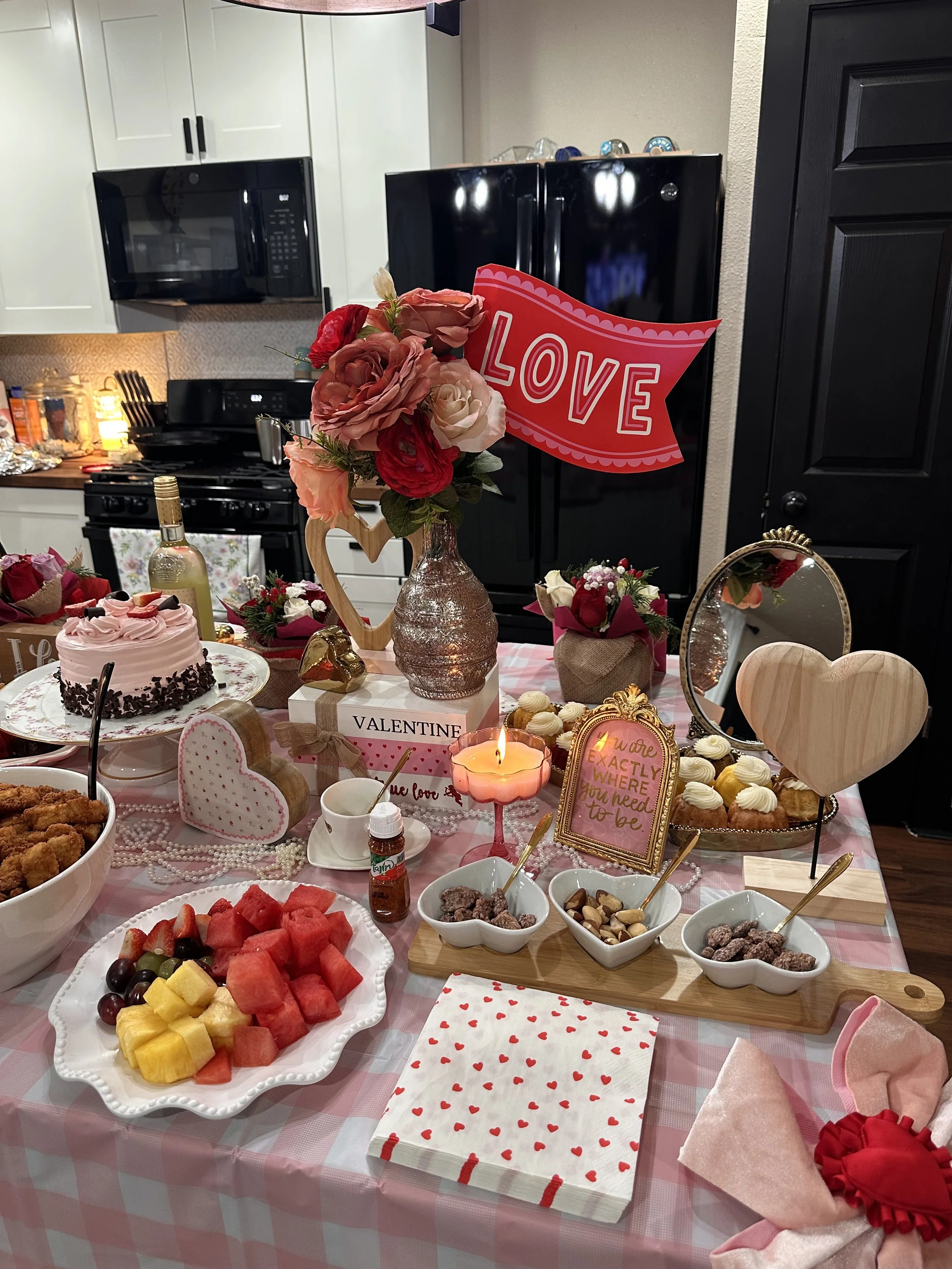 A table decorated for Valentine's Day with assorted desserts and fruit. There are pink and red flowers, a sign that says 'LOVE,' and small hearts and romantic-themed items. The table has a pink checkered tablecloth and various serving dishes with tre