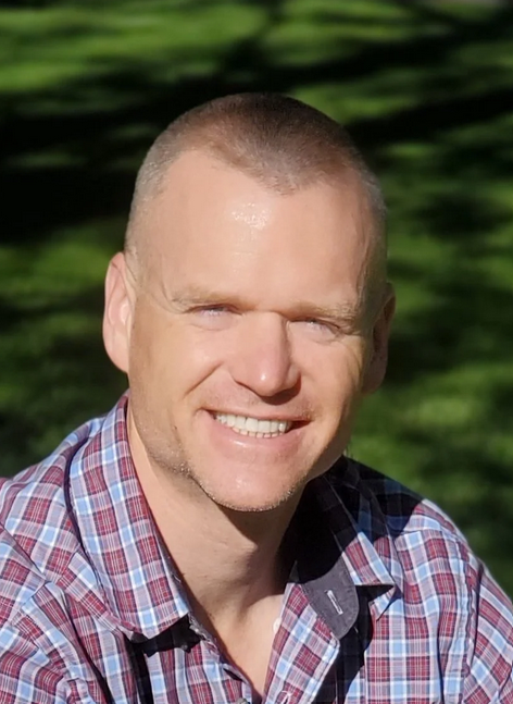 A smiling man with short hair wearing a plaid shirt outdoors with green grass in the background.